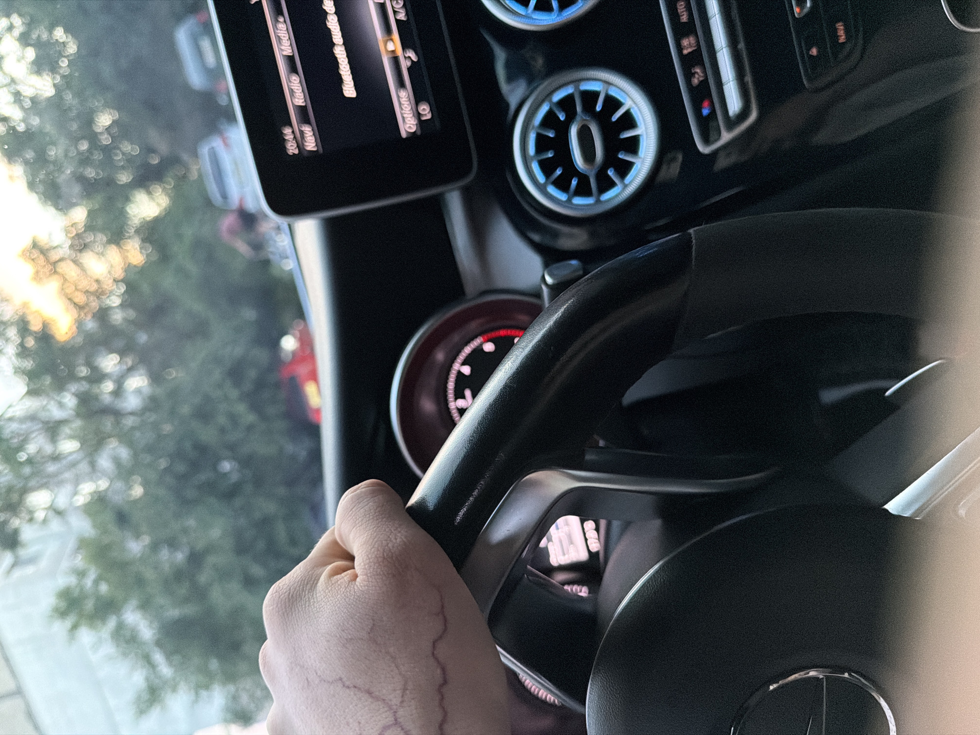 The image presents a first-person view from inside a car, identified as a Mercedes-Benz by the partial logo on the steering wheel, in Shumen, Bulgaria. A person's right hand, showing prominent veins, is gripping the black steering wheel. The car's dashboard features circular air vents with a distinctive blue backlighting and a section of the instrument cluster glowing with red light.
An infotainment screen is visible above the dashboard, displaying several menu options. Visible text on the screen includes "Navi," "Radio," "Media," "Options," and "Bluetooth audio de." The time "20:41" is clearly shown next to "Navi," indicating an evening time. Below "Options," the letters "LO" are visible. Additionally, "A/C" and "AUTO" are partially visible on adjacent control panels.
Through the driver's side window, a blurred background of green foliage and warm-toned, out-of-focus light sources is visible, suggesting either direct sunlight or strong artificial light in the evening. The scene depicts an individual either driving or stationary in the car during the evening hours.