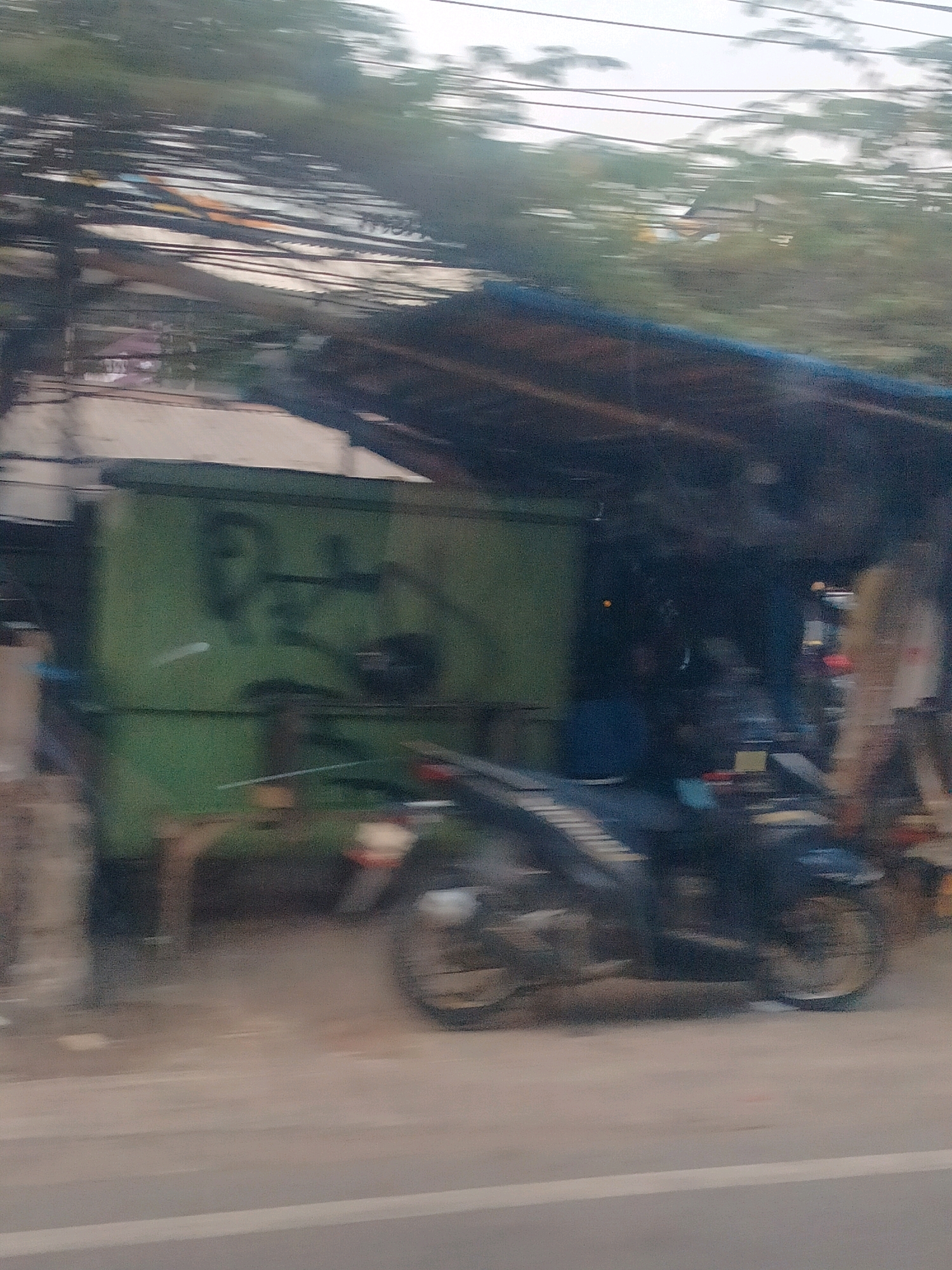 This is a motion-blurred image taken from a vehicle, likely a car or bus, driving on a street in East Jakarta, Indonesia. The scene captures a roadside stall or structure with a green wall, possibly a container. The green wall has some dark graffiti on it. Above the stall, a dense network of electrical wires is visible against a light sky. In front of the stall, a dark-colored motorcycle is parked or moving, with its wheels and part of its body clearly visible. To the right of the motorcycle and stall, a person in a light-colored shirt is partially visible, standing near what appears to be more stall structures. The overall impression is a busy, slightly cluttered urban environment typical of a roadside setting in Indonesia.