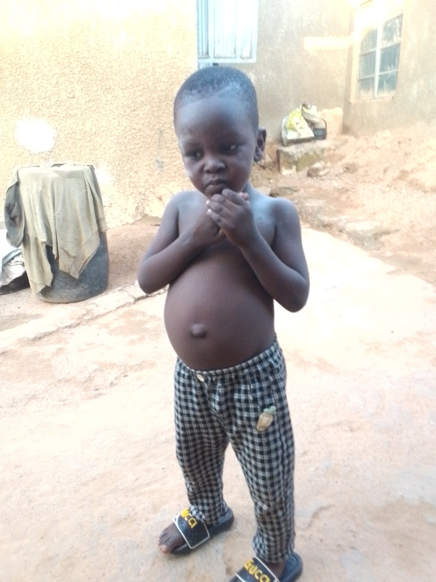 A young child, appearing to be around three years old, stands on a dirt-like surface in front of a textured, beige-colored building. The child is shirtless, revealing a distended abdomen, and wears black and white checkered trousers and black slides with a yellow logo. The child's hands are raised to their mouth, and their expression is contemplative. To the left of the child, a large, dark cylindrical object, possibly a barrel, is covered with a light-colored fabric. In the background, a window with white shutters is visible on the building, and to the right, there is a pile of objects, including what appears to be a bag with yellow and white contents. The ground is uneven and appears to be dry earth or sand. The lighting suggests it is daytime. The location is identified as Narayi, Nigeria.