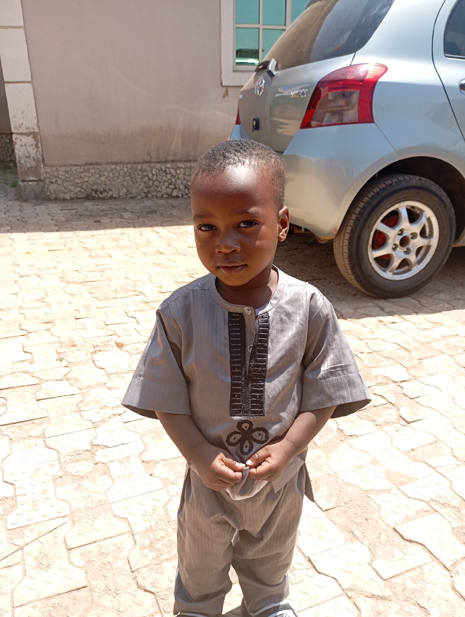 A young child stands outdoors on a paved surface composed of interlocking, light-colored concrete blocks. The child is wearing a gray, short-sleeved tunic-style outfit with dark embroidery along the chest. To the right and slightly behind the child is a silver Toyota Yaris parked close to a building with a window. The building is light beige with a darker stone base. The scene appears to be in Jos, Nigeria, based on the background elements and the ethnicity of the child.