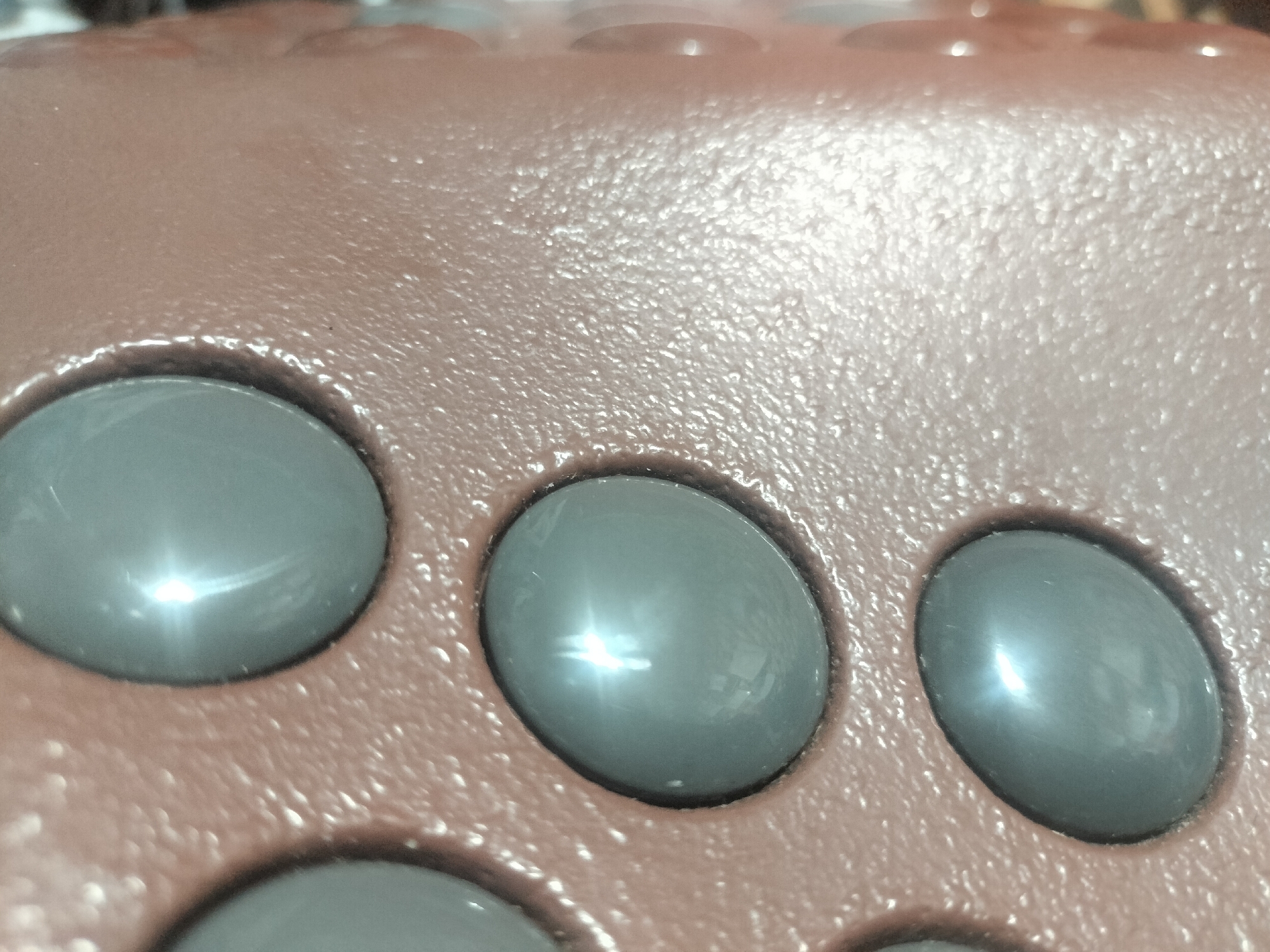 The image shows a close-up, eye-level shot of a brown, textured surface with several circular, dark gray indentations. The indentations appear to be part of a larger object, possibly a game board or a piece of equipment. Light reflects brightly off the surfaces of these indentations, creating starburst-like highlights. The brown surface has a slightly bumpy texture, visible in the spaces between the indentations. The background is out of focus and appears to be a continuation of the same brown material, with more indentations faintly visible. There are no people or animals in the image, and the setting is not clearly identifiable as being in Akbasheva, Russia, or any other specific location. The image focuses solely on the texture and form of the object.