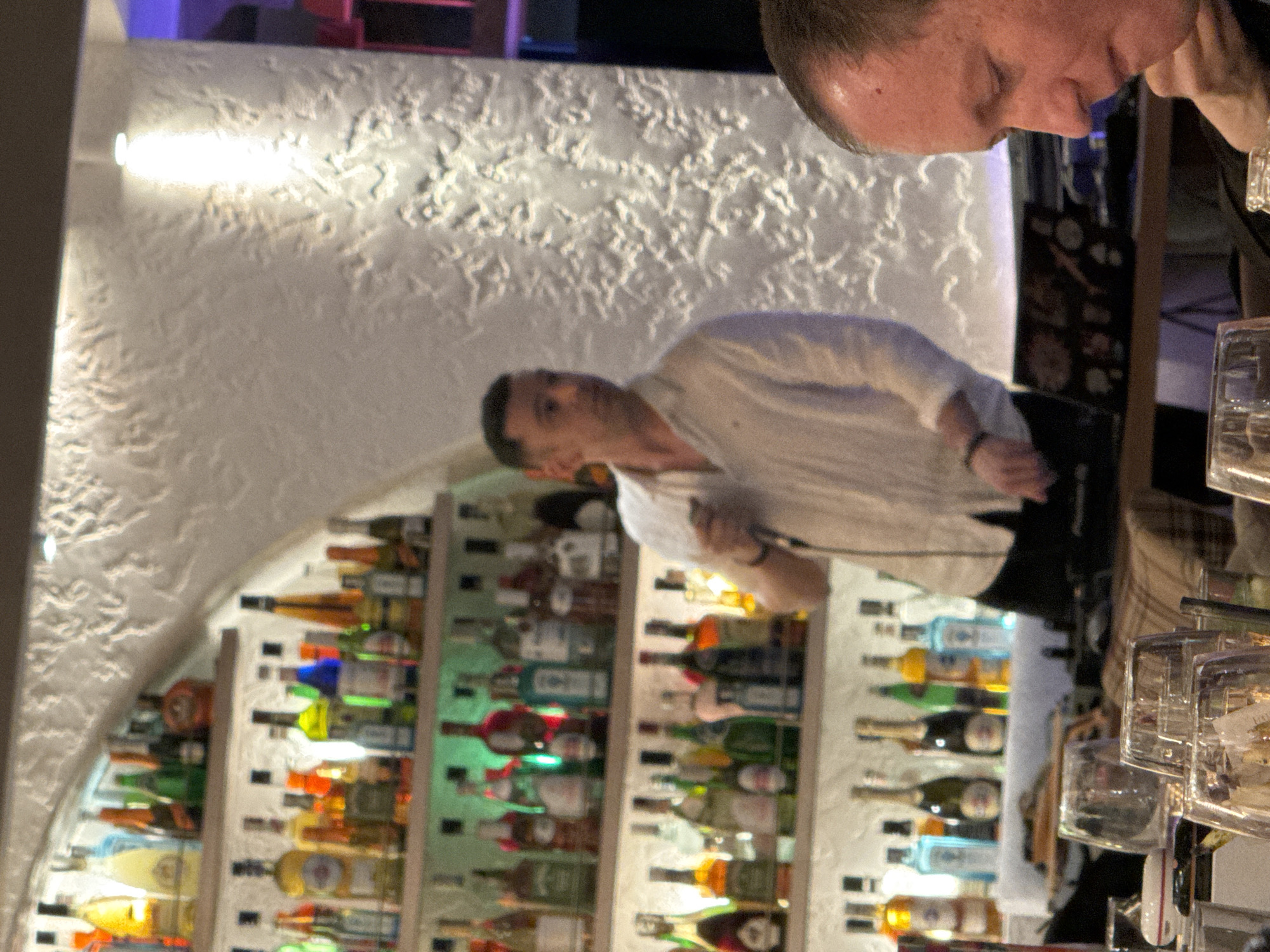 A sideways shot shows two men standing at a well-stocked bar, seemingly in a nightlife establishment. A man in a white, long-sleeved shirt is positioned in the center, with a concerned expression on his face. Another man, with short hair, is standing beside him with his head turned, appearing to engage in a transaction or a conversation with the bartender. Behind the men, shelves of assorted bottles are lit up, creating a colorful backdrop. The location appears to be in a bar within the city of Sofia, Bulgaria.