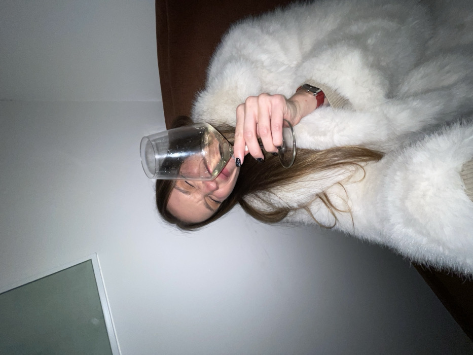 A woman in a fluffy white coat reclines on a brown surface. She holds a glass of liquid to her mouth, her eyes closed in apparent discomfort. Her dark hair falls beside the glass. A red-banded wristwatch is visible on her wrist. The background includes a white wall with a window or mirror frame in the lower left corner. The image is shot at an unusual angle.