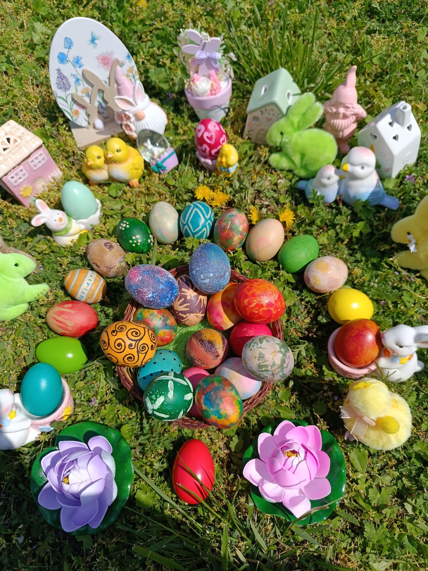 The image depicts an Easter scene set in a grassy area, potentially in Cherkovitsa, Bulgaria. The arrangement includes a basket filled with painted Easter eggs, surrounded by individual eggs placed on the grass or in decorative holders. Various Easter-themed figurines and decorations such as chicks, rabbits, miniature houses, gnomes, and flowers are scattered around the eggs. The scene is bathed in sunlight, suggesting an outdoor setting.