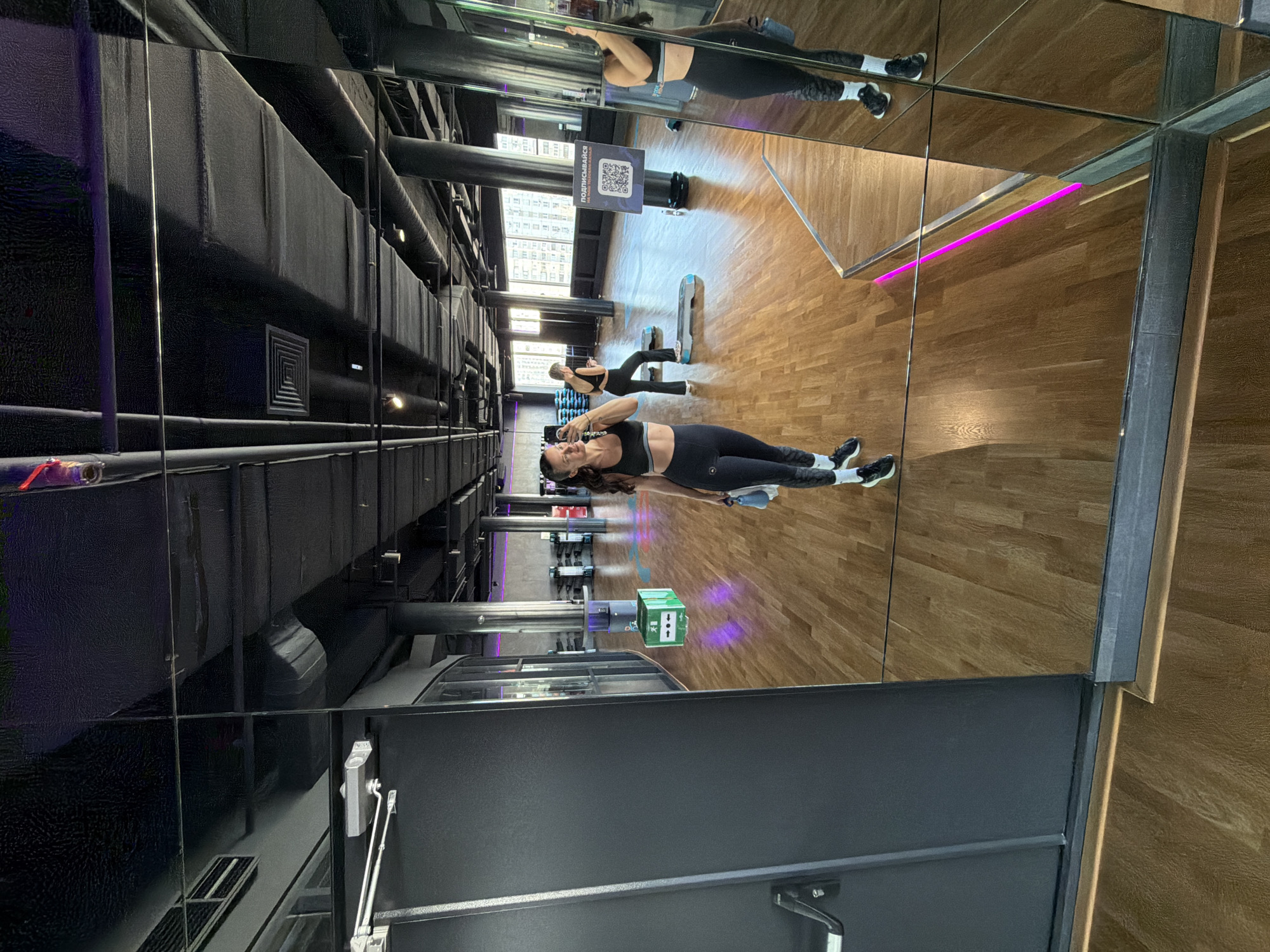 The image depicts the interior of a modern gym, observed through a large mirror. The facility is located in Moscow, Russia, as indicated by Cyrillic text on signs within the scene; the precise street address or name of the gym is not visually identifiable.
The environment features a light-colored wooden floor and a high, dark gray/black industrial-style ceiling with exposed ventilation ducts, pipes, and recessed lighting fixtures. Dark gray structural columns are distributed throughout the space. Large windows line one wall, providing natural light and a view of an urban cityscape with multi-story buildings in the distance. Accent lighting includes purple LED strips along floor edges and possibly architectural features.
The primary subject is a female with long, dark, wavy hair, visible in the foreground as a reflection in the mirror. She is smiling at the camera (a smartphone held in her right hand, obscuring part of her face). She wears a black athletic crop top, black full-length leggings with a subtle gray pattern on the lower legs, white ankle socks, and black athletic shoes. In her left hand, she holds a light blue water bottle. Her action is taking a mirror selfie.
A secondary female subject is visible in the midground, further back in the reflection. She wears a black sleeveless top and black flared athletic pants. She stands near a gray aerobic step platform, looking down at a smartphone in her hands, indicating an interaction with her device rather than active exercise.
Objects and equipment present include multiple gray aerobic step platforms on the floor, a freestanding water cooler dispenser with a blue bottle, and a green storage box with white arrows and Russian text. A tall, black rectangular sign displays a white QR code and Russian text reading "ПОДПИСЫВАЙТЕСЬ НА НАШИ СОЦСЕТИ" (Subscribe to our social networks) and "ОБЯЗАТЕЛЬНО ПРОЙДИТЕ ОНЛАЙН-РЕГИСТРАЦИЮ" (Mandatory online registration). A dark gray door frame with a metallic door closer mechanism is visible on the left side of the frame. There are no direct interactions between the two visible individuals.