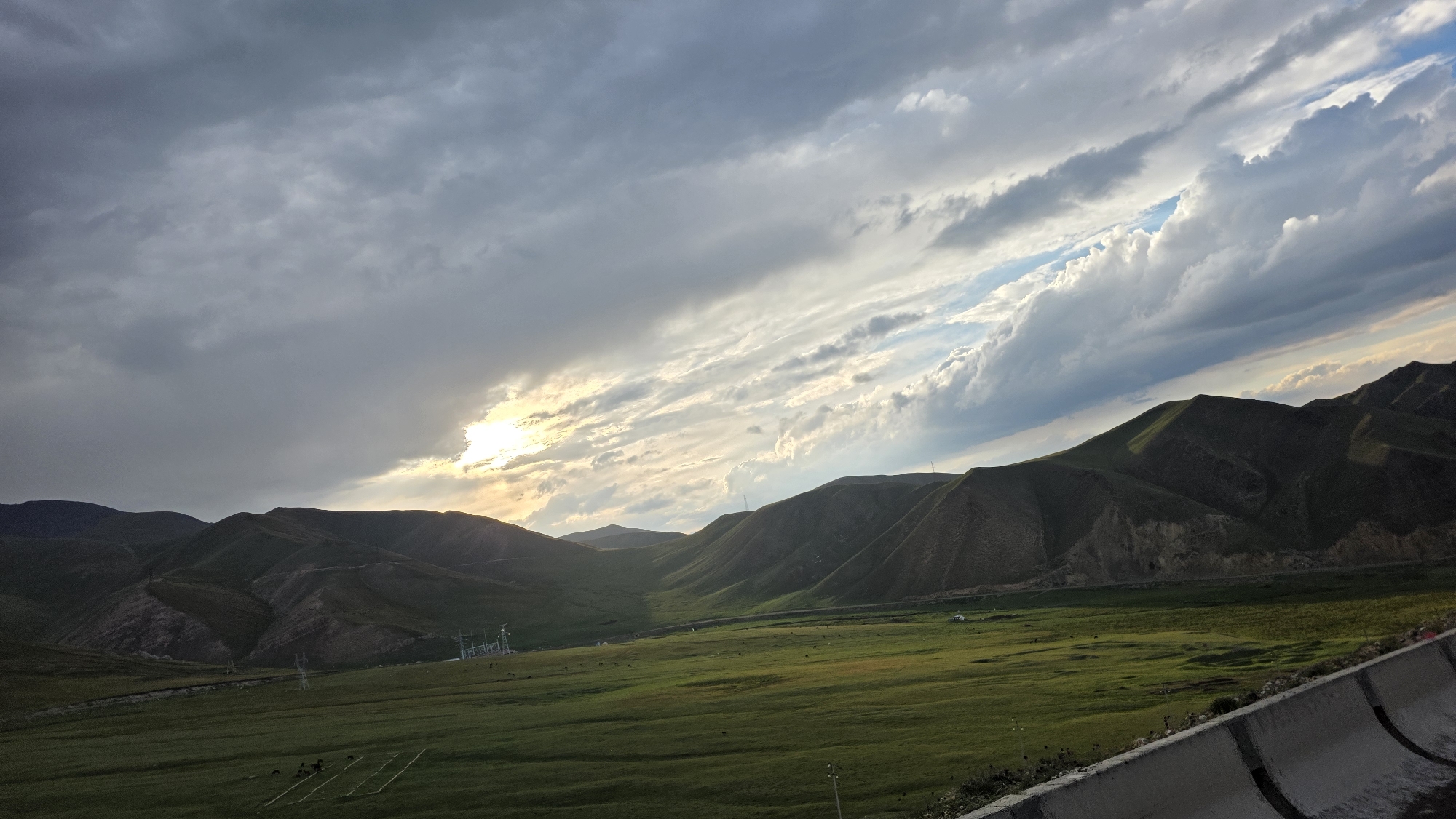 A wide shot captures a landscape in Kara-Unkur, Kyrgyzstan, under a dramatic sky. Rolling green hills dominate the middle ground, with some showing exposed rock faces. A flat, green grassy plain stretches between the hills. Power lines and towers are visible on the left side of the plain, with a few small structures and what appear to be livestock scattered across the field. A concrete barrier runs along the bottom right edge of the frame, suggesting a road or overlook. The sky is filled with a mix of dark, heavy clouds and streaks of bright, wispy clouds, with the sun breaking through on the left, casting a warm glow on the scene. The overall impression is one of rugged natural beauty with signs of human infrastructure.