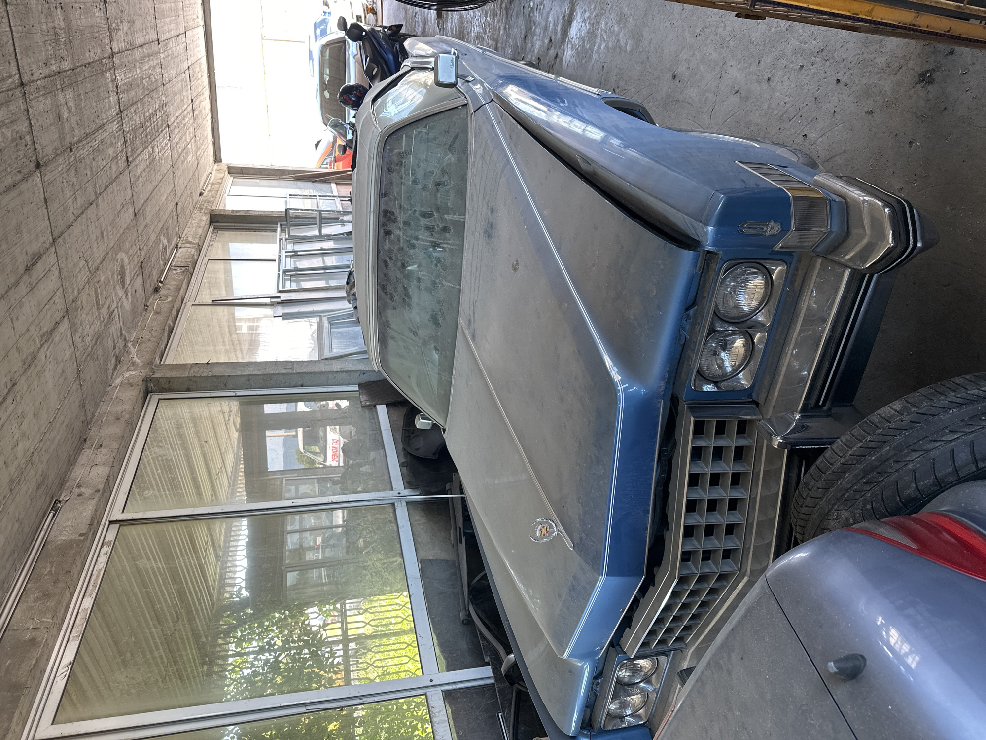 An older, dusty, light blue Cadillac Eldorado convertible is parked indoors, seemingly in a garage or storage unit. The car is the primary subject, positioned prominently in the foreground and extending into the midground. Its hood ornament, a chrome Cadillac crest, is visible. The car has a large, squared-off grille and dual headlights.
To the left of the Cadillac, a portion of a gray car is visible, with a tire partially obscuring the Cadillac's front bumper. In the background, other vehicles are present, including a silver sedan and a red car. The environment appears to be a somewhat disused indoor space with a concrete ceiling and walls, featuring large windows that allow natural light to filter in, revealing greenery and a white van with text on its side visible through the glass. Stacked window frames are also visible against the wall. The overall impression is one of a collection of stored vehicles in a utilitarian setting.