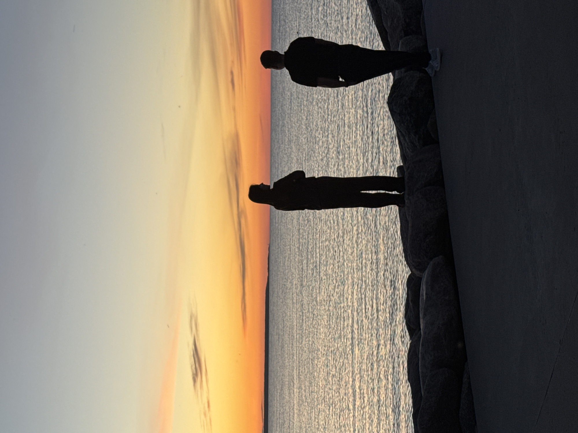 Two individuals, silhouetted against a sunset, stand on a rocky shoreline in Tallinn, Estonia. The person on the left is facing left with their arms crossed, while the person on the right is facing the sea. The sky displays a gradient of colors, from pale blue at the top to a vibrant orange near the horizon, with wispy clouds. The sea is calm, reflecting the sky's colors. A paved walkway is visible in the foreground. The overall scene depicts a serene evening at the coast.