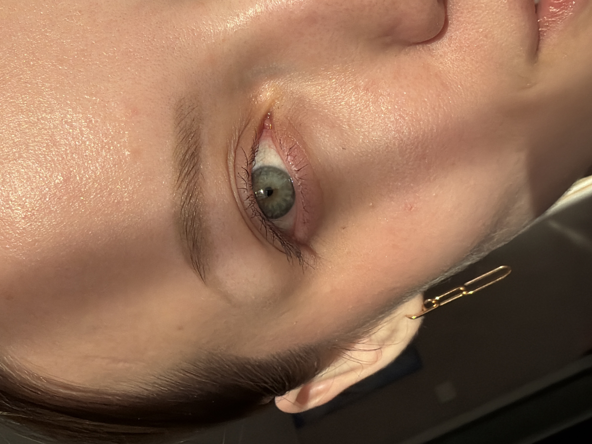 A close-up, rotated view of a person's face focusing on their left eye. The eye is open, with gray-green irises and dark eyelashes. The skin appears smooth with some subtle textures. A gold-colored earring in the shape of a linked paper clip is visible on the left side of the image, attached to the ear. The background is dark and indistinct. There is no information given or visually discernible to identify the location as Reshetnikovo, Russia.