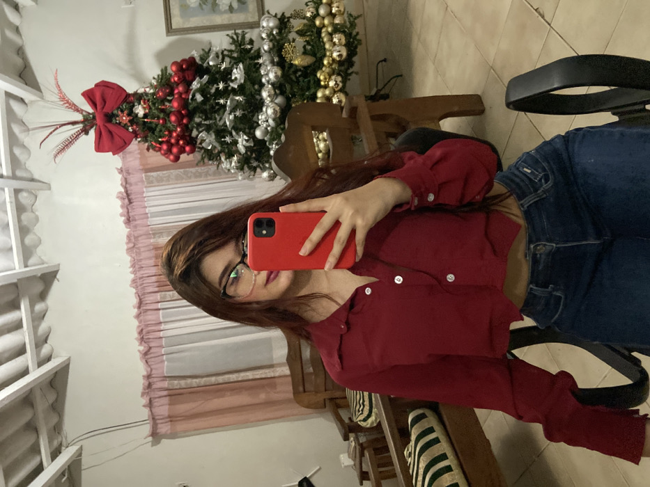 A young woman in a red button-up shirt and blue jeans sits in a black office chair, taking a selfie with a red phone. Behind her, a decorated Christmas tree stands near a window with pink curtains. The room features light-colored walls, a wooden chair, and glimpses of the ceiling's white wooden structure. Her brown hair partially covers her face, and she wears glasses.