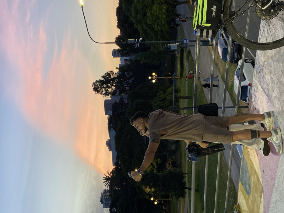 A man takes a photo with his phone. He wears a brown hooded shirt and shorts with white shoes. A bike is parked nearby with a food delivery bag attached. Behind the man is a street scene with a grassy area and trees. Buildings are visible in the background and the sky features a blend of blue and orange hues, suggesting either sunrise or sunset.