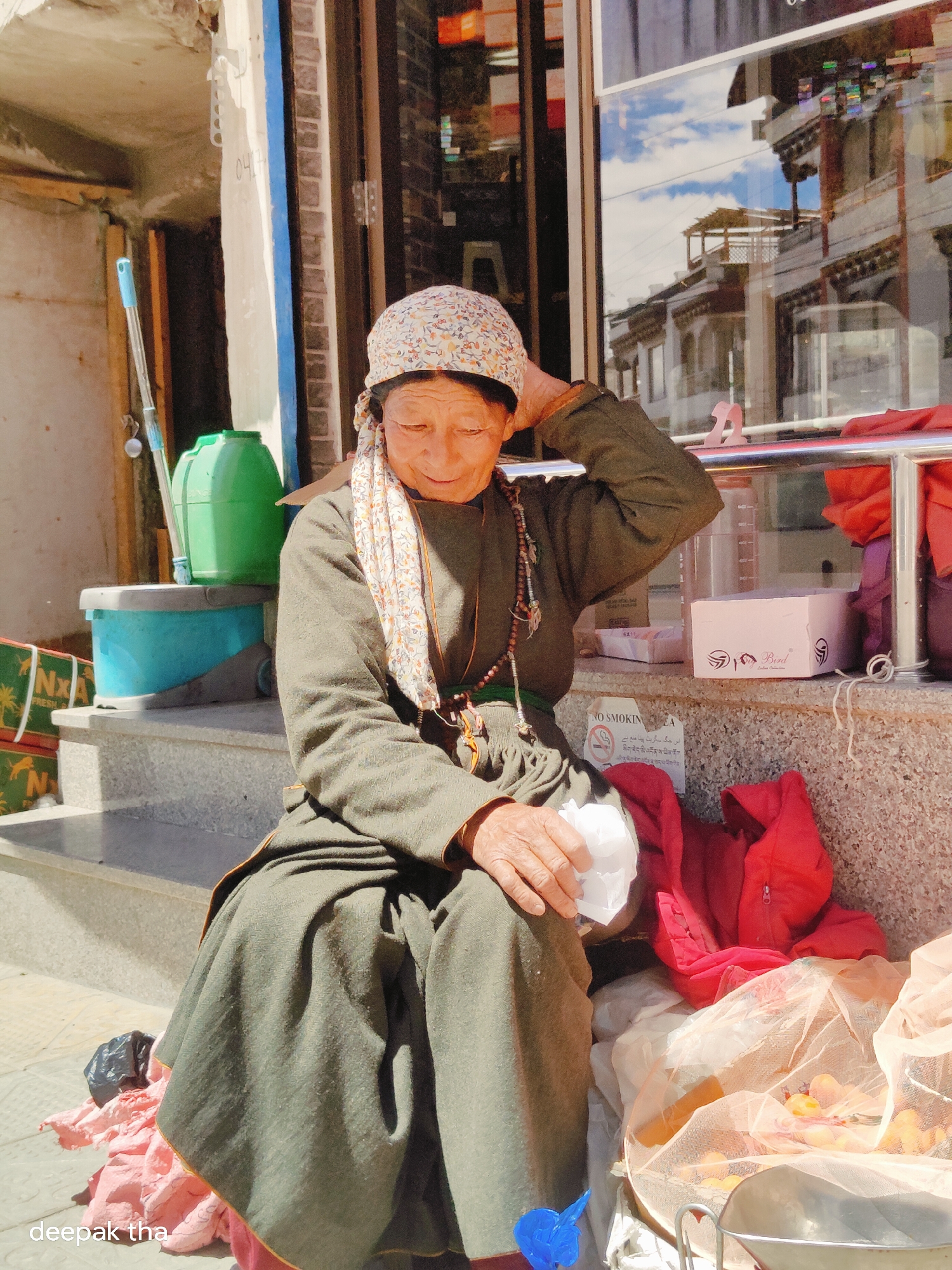 An elderly Tibetan woman with a patterned headscarf and a floral scarf around her neck sits on concrete steps in Leh, India. She wears a dark olive-green traditional garment. Her right hand is raised to her head, and she holds a white tissue in her left hand. She is adorned with a beaded necklace. To her right, a pile of red clothing and a mesh bag containing fruits are visible. In the background, a shop window reflects the sky and buildings. To her left, a green plastic container and a blue bucket are placed on the steps. The scene appears to be on a street in Leh.