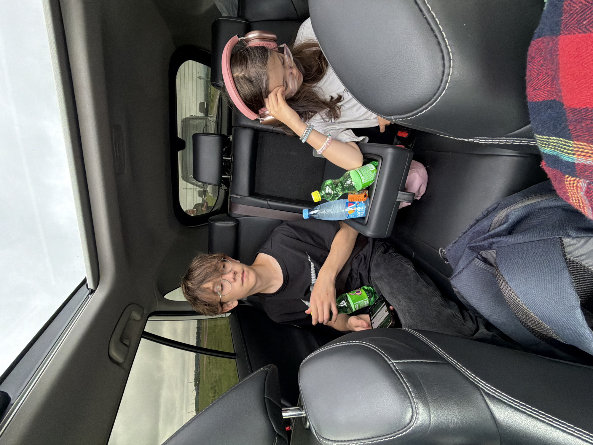 The image shows the interior of a car. Two young people, a boy and a girl, are seated in the back. The boy, wearing glasses and a black t-shirt, is holding a green bottle and a smartphone. The girl, wearing pink headphones and glasses, has her hand on her head. There are two bottles of soda in a cup holder between them. Outside the car, a field and a cloudy sky are visible, suggesting a rural or suburban location, possibly near Кирга, Russia. The overall scene depicts a moment of travel or rest during a journey.