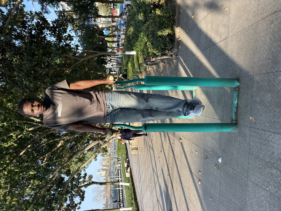 The image depicts a daytime outdoor scene within a public park or promenade situated along a riverfront in Jiangjin, China.
A Black adult male, approximately 20-30 years old, stands centrally positioned on a teal-colored outdoor air walker exercise machine. He is wearing a dark grey short-sleeved t-shirt with "MOCOME" printed on the chest, light blue jeans, and grey athletic shoes with white soles. His hands grip the yellow spherical handles of the equipment, and his feet rest on the suspended foot platforms. He faces directly towards the camera with a neutral expression. Sunlight casts distinct shadows across his torso and legs. The exercise machine exhibits signs of wear, including chipped paint and rust at its base.
The ground consists of grey square paving tiles with visible grid lines, scattered with a few brown leaves and a small piece of white debris. Long, angular shadows from overhead trees extend across the pavement from the right towards the left.
In the midground to the left, a paved walkway continues, bordered by green grass and a low stone or concrete barrier. Beyond this, a wide body of water, identifiable as the Yangtze River, is visible, with the distant opposite bank featuring a dense cluster of multi-story urban buildings under a clear blue sky. An East Asian male, dressed in a dark jacket, is walking away from the camera on the left walkway, and another distant figure is partially visible near the river.
To the right, a landscaped area with green hedges and numerous trees with green foliage runs alongside the path. A black bicycle is parked upright near the hedges. Further beyond the trees, an urban street or boulevard is discernible, showing vehicles (possibly buses) and additional buildings, indicating a continuous urban environment. The scene is illuminated by strong, direct sunlight.