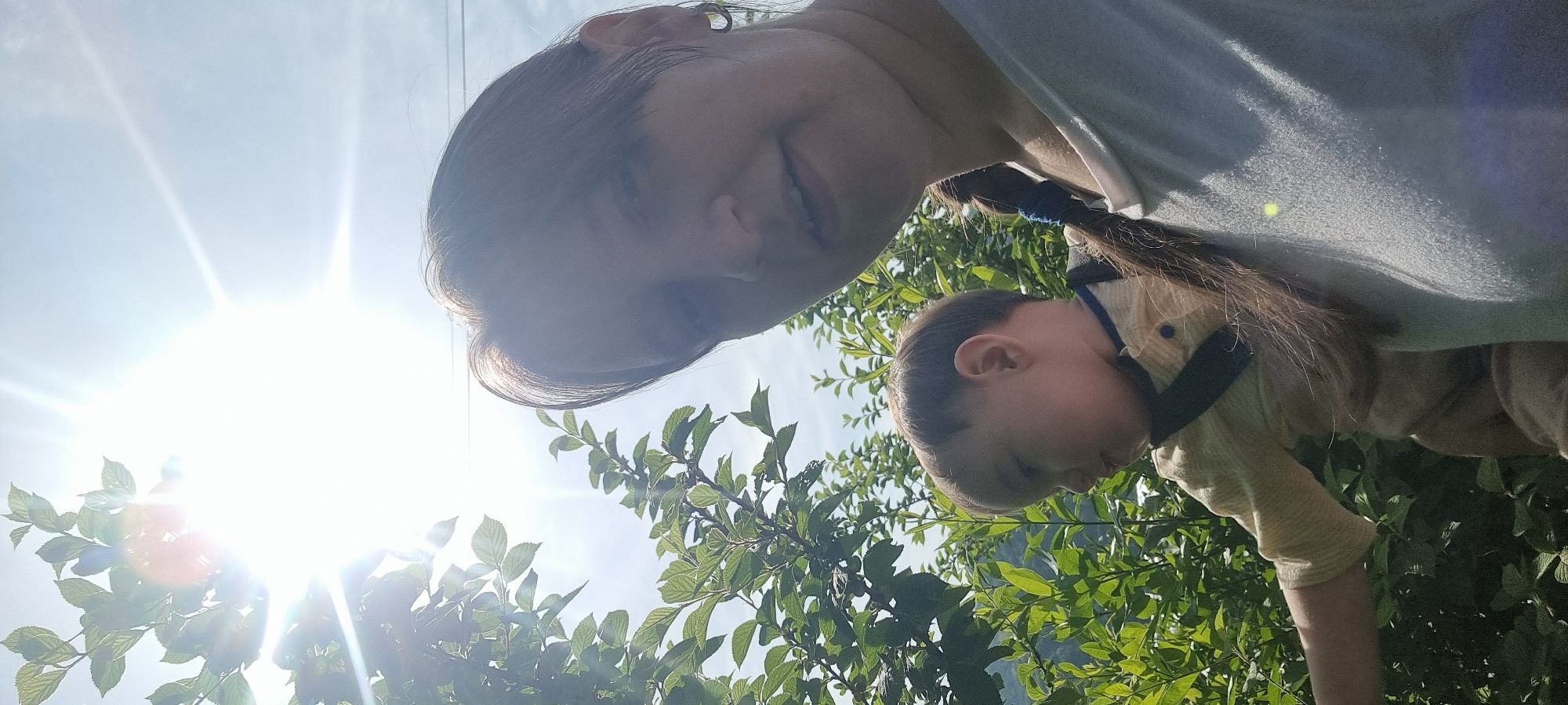 A woman and child stand outdoors in Dunnae-myeon, South Korea. The sun is high, creating bright light. Green trees and foliage fill the lower portion of the frame. The woman is fair-skinned with brown hair in a braid. The child is fair-skinned with light brown hair, wearing a yellow shirt.