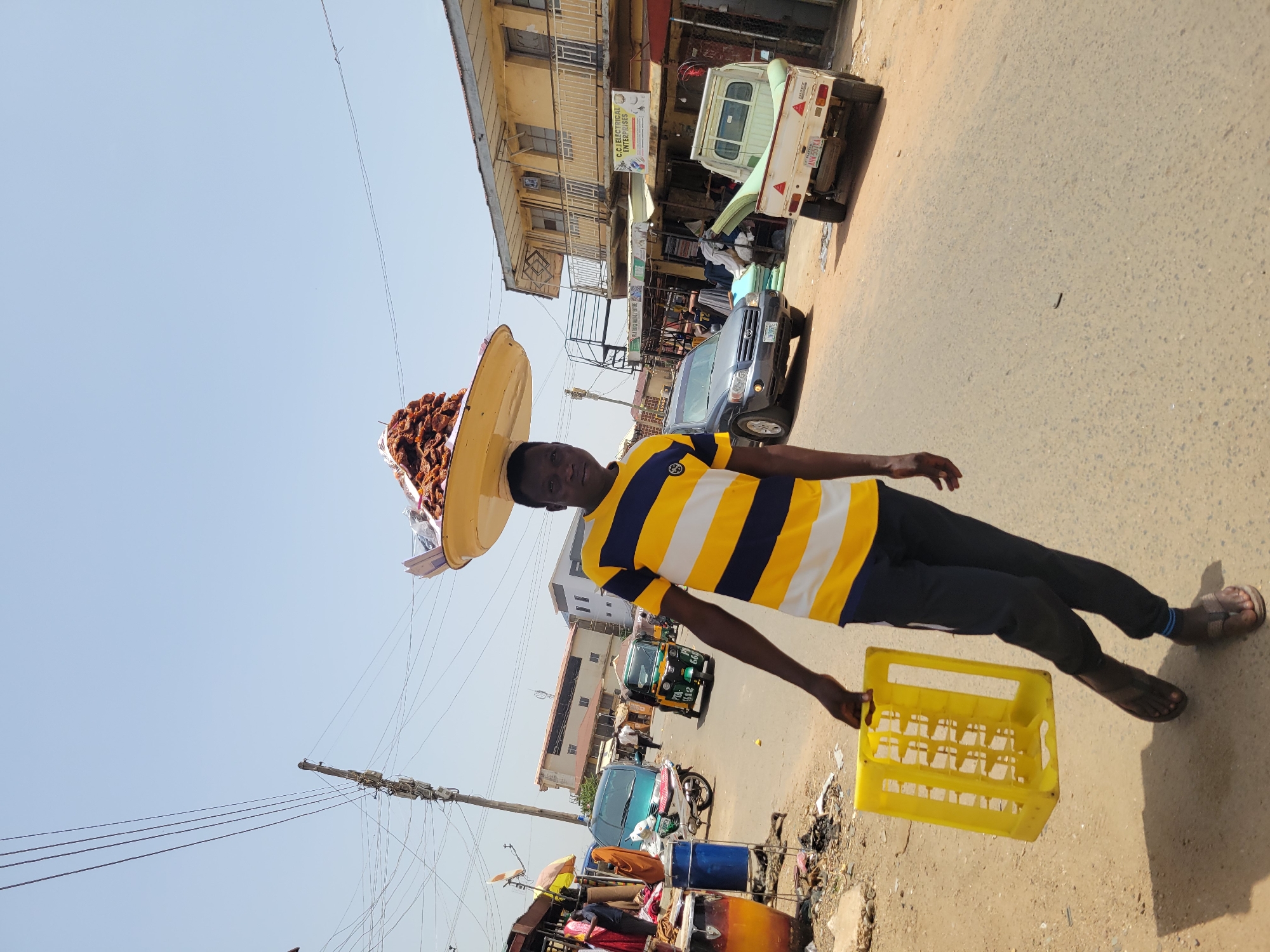 In a street scene in Jos, Nigeria, a young man balances a large, round tray loaded with brown objects on his head. He is wearing a horizontally striped yellow and navy blue shirt, dark pants, and sandals. In his hand, he carries a yellow plastic crate. He stands on a paved road with buildings visible in the background. The road shows signs of wear and tear. Various vehicles, including cars and auto-rickshaws, are parked or moving along the street. Debris and waste are scattered along the side of the road where the man is standing. Overhead power lines are visible against a clear sky. The photograph captures a typical urban street scene.