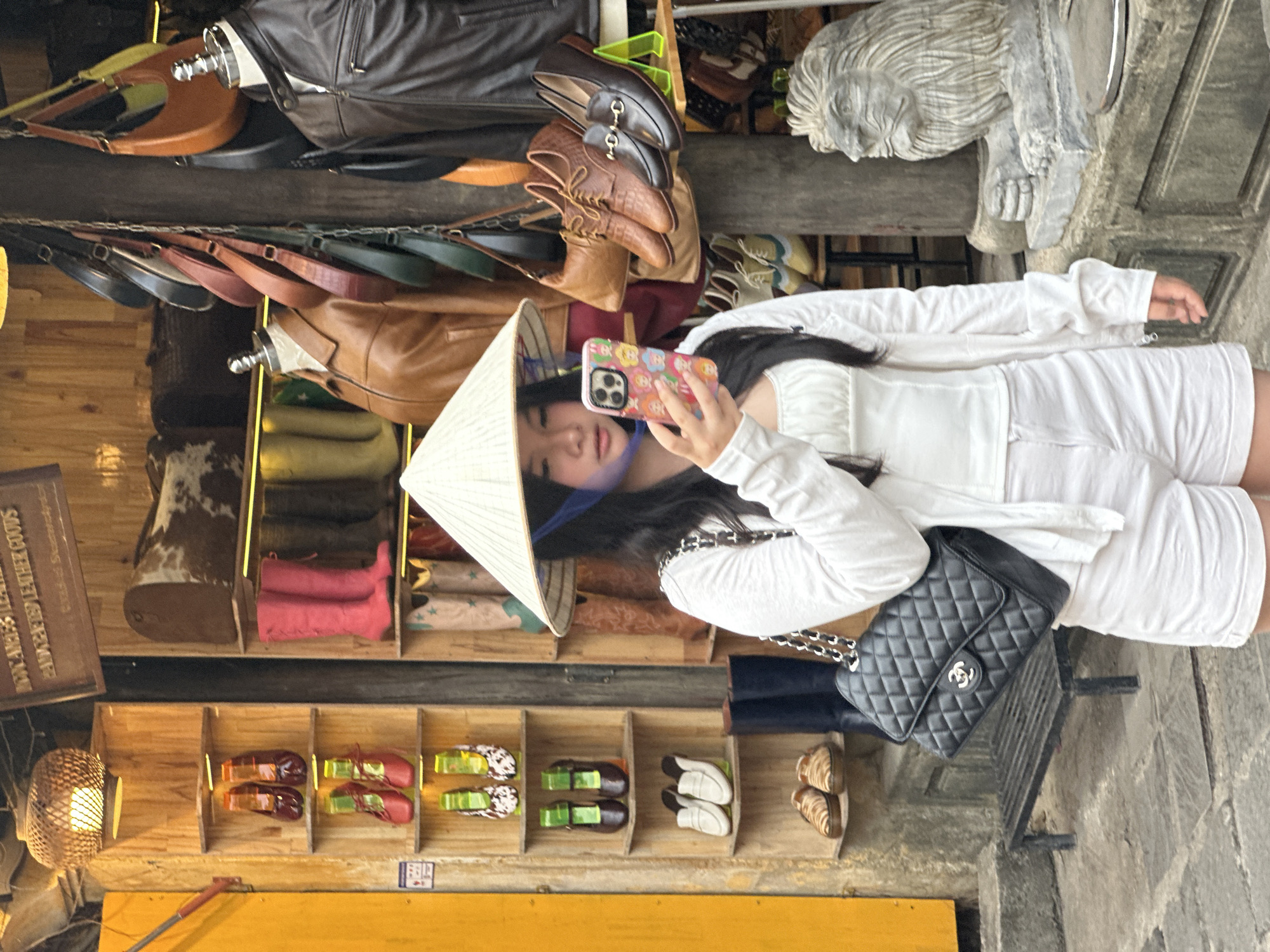 In Hội An, Vietnam, a young woman, wearing a conical hat, takes a selfie with her phone. She is dressed in an all-white outfit, comprising a white long-sleeved top and white shorts. A black quilted handbag with a silver chain strap is slung over her shoulder. The background suggests a market or shop, with various leather goods, including bags and shoes, displayed on shelves. Wooden shelves are filled with items like boots and small decorative shoes. A sign with faded text is visible on the left, and a woven lamp hangs from the ceiling. The scene appears to be outdoors, possibly in the daytime, given the lighting.