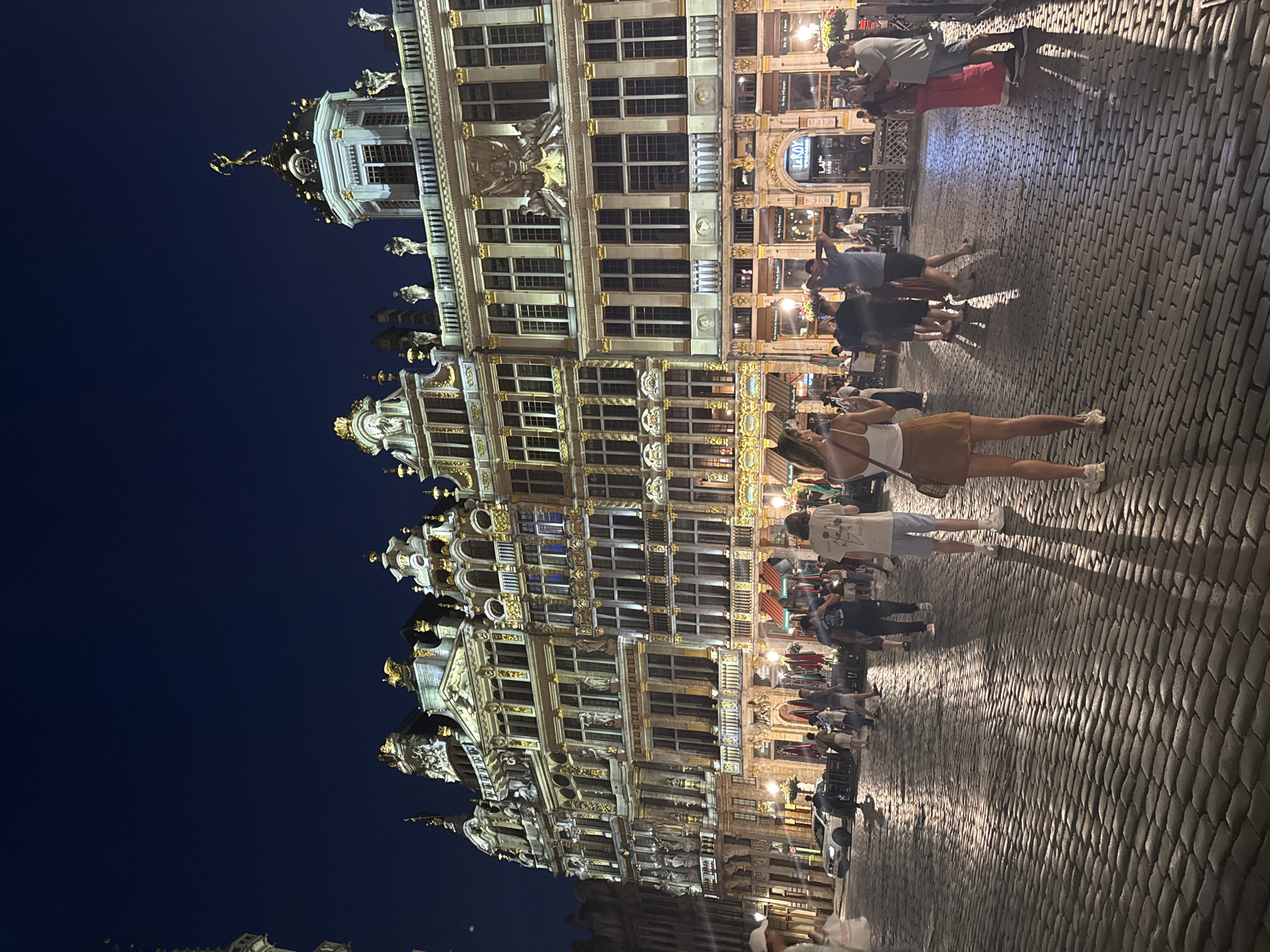 The image depicts the Grand Place in Brussels, Belgium, at night. Ornate, gilded buildings with intricate architectural details line the cobblestone square. Several people are present, walking and standing. In the foreground, a woman with blonde hair wearing a white tank top and a brown skirt is taking a photograph with her phone. Other individuals are scattered throughout the square, some appearing to be tourists. Shadows are cast across the illuminated cobblestones, indicating the direction of the lighting. The sky is a deep, dark blue.