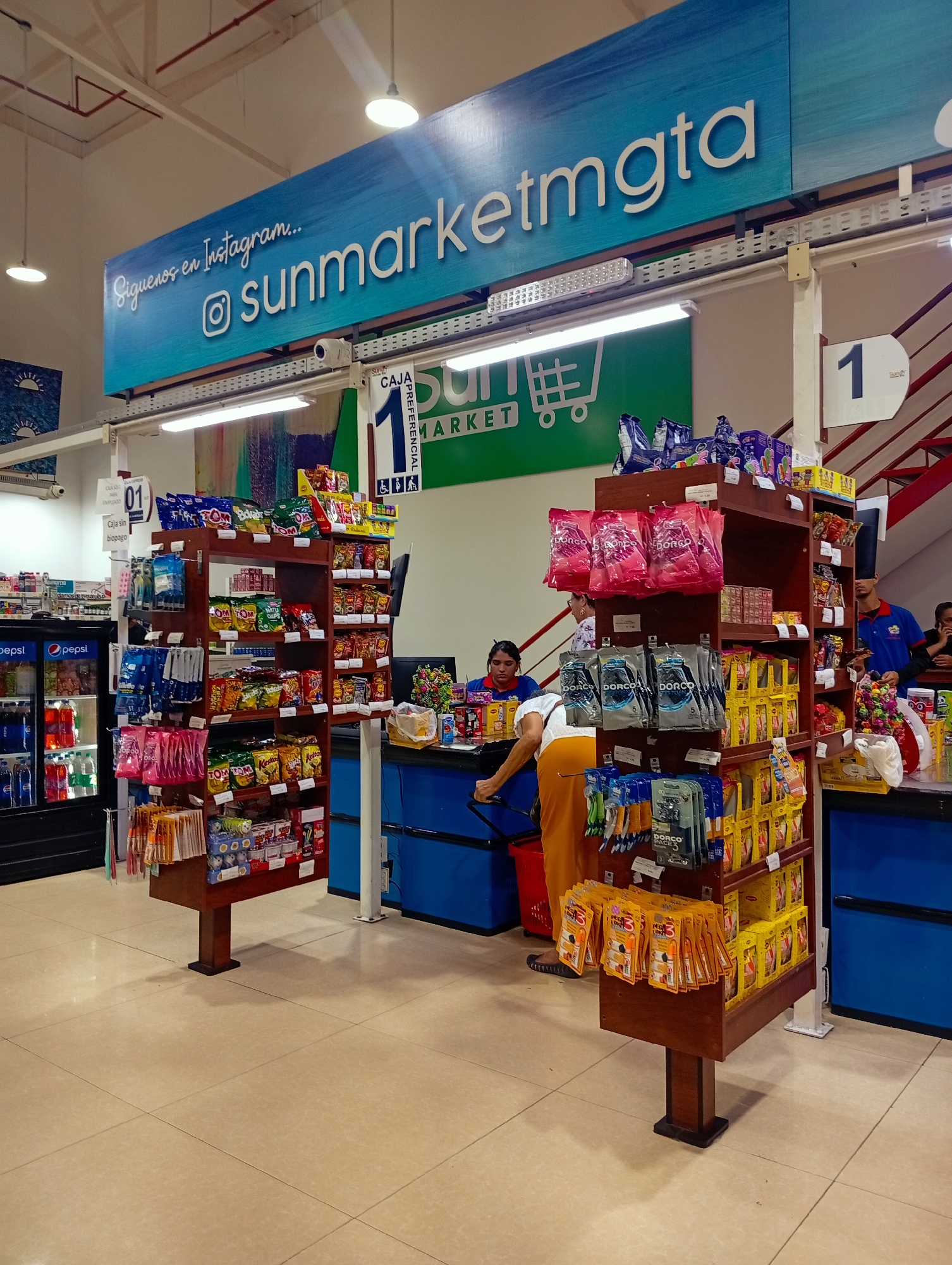 Here is a detailed description of the image:
**General Description:**
The image captures the interior of a Sun Market supermarket, specifically at the checkout area. It appears to be a typical day, with customers and employees present. A sign at the top says “Siguenos en Instagram” and mentions the name of this store “sunmarketmgta”.
**Setting and Environment:**
The setting is the checkout section of a supermarket. There are tile floors and high ceilings. The checkout lanes are numbered. Bright lighting is present.
**People:**
There are multiple people visible, including:
* Cashiers working behind the counters.
* A customer with mustard colored pants making a purchase.
* A male worker in a blue shirt stands near the checkout lanes.
**Objects:**
* Checkout counters with blue cabinets.
* Candy and snack displays flanking the counters.
* Refrigerated display case with Pepsi products.
* Various groceries and products on shelves.
* Checkout lane dividers
**Actions and Interactions:**
* Cashiers are actively serving customers at the checkout counters.
* Customers are going through the checkout process, purchasing items.
**Inferred Information:**
The image suggests a typical shopping experience at a local supermarket. It offers a glimpse into the daily activities within a business establishment in Venezuela.
Based on the presence of the Sun Market logo and the sign mentioning Instagram with the handle "@sunmarketmgta," and considering the question's request for location information, it's logical to conclude the image was taken at a Sun Market supermarket in Juan Griego, Venezuela, though this cannot be definitively confirmed from the image alone.