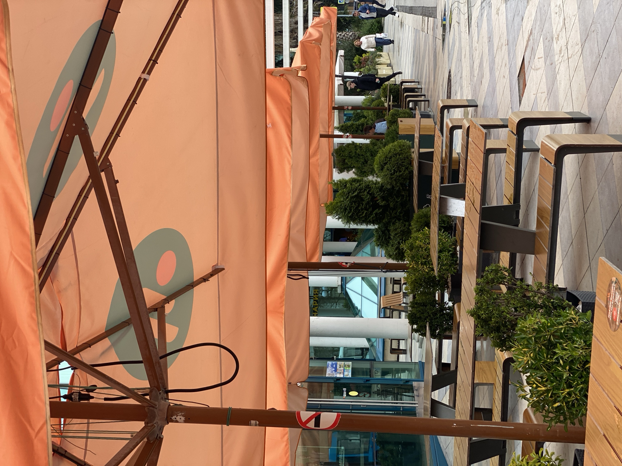The image shows an outdoor cafe in Sochi, Russia. Tables and benches line a pedestrian walkway, separated by small green trees in black planters. Each table has a large peach-colored umbrella with a blue and pink graphic. Several people walk in the distance on the tiled walkway. The background includes a building with glass windows and white pillars. A sticker with the number "1" inside a red prohibition symbol is on the umbrella.