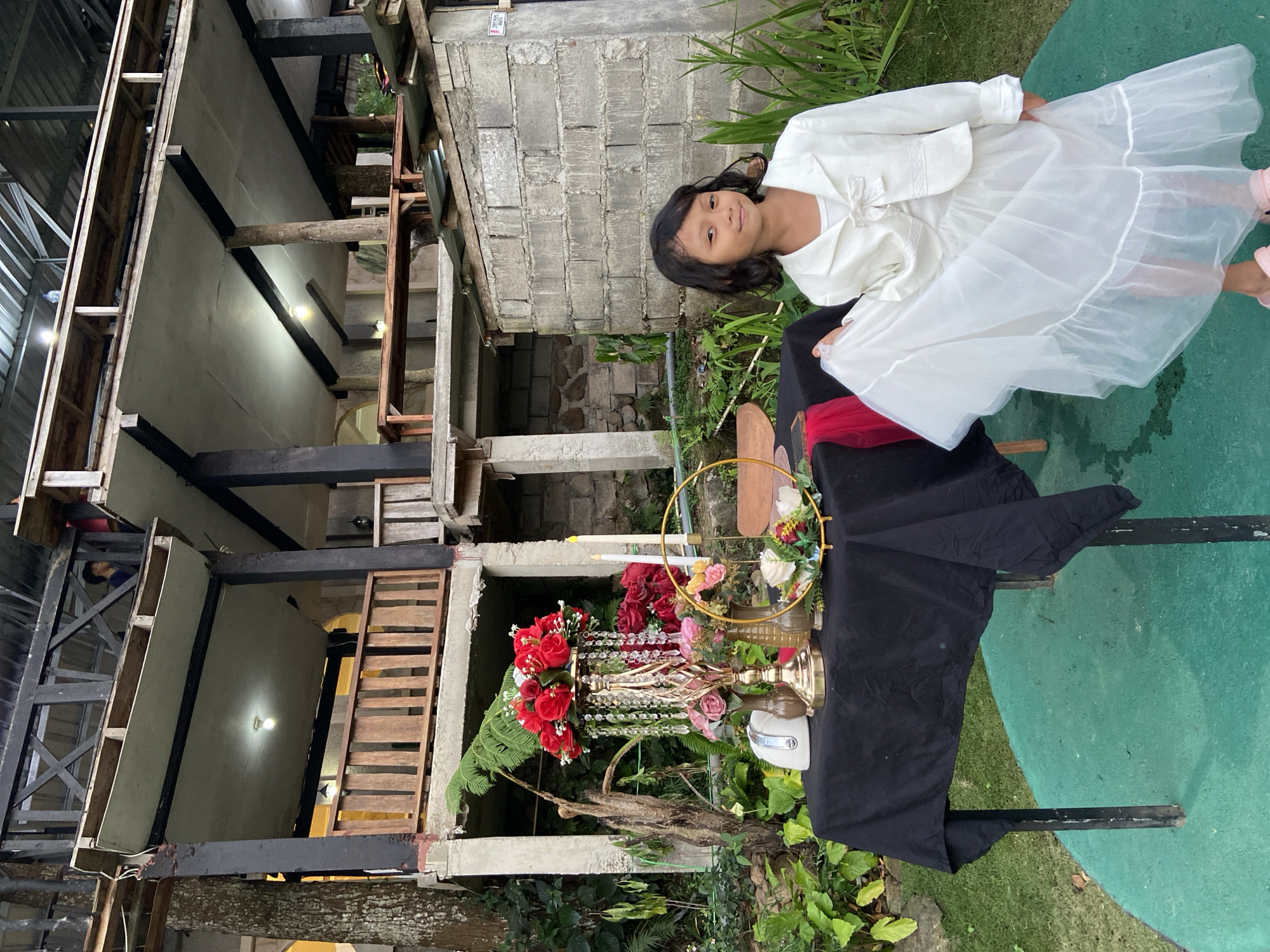 In the city of Kesiman, Indonesia, a young girl stands next to a table adorned with flowers and a black tablecloth. She is wearing a white jacket and a white dress. The table is set with floral arrangements, candles, and decorative items. In the background, a multi-story building with wooden structures is visible, along with lush greenery. The setting appears to be outdoors, possibly a garden or a venue for an event.