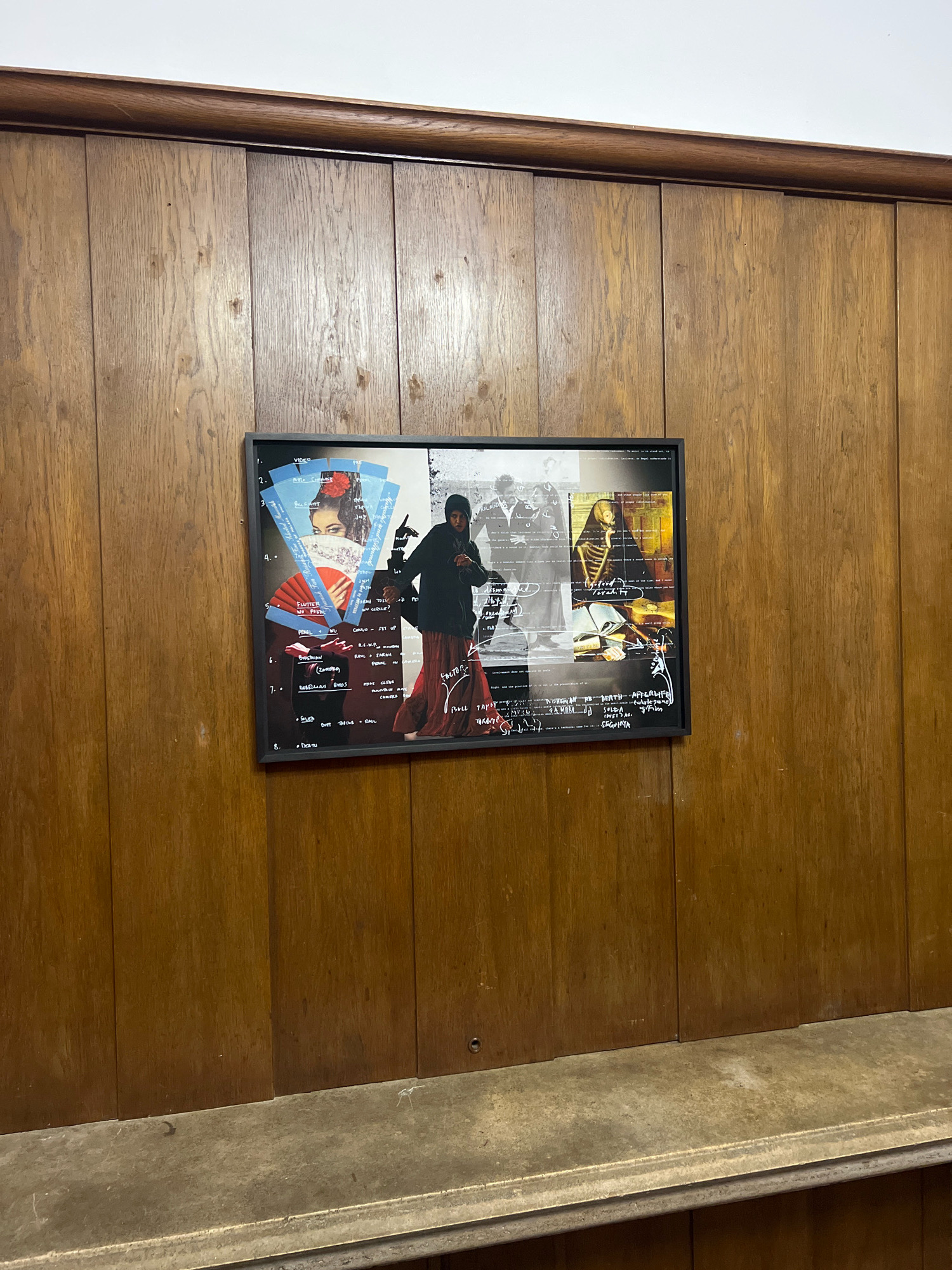 This is a medium shot, eye-level photograph of a framed collage of images and text hanging on a wood-paneled wall. The collage features multiple photographic elements, including a woman wearing a red fan and a black veil, a silhouetted figure in a red dress, and a skeleton. There is also handwritten text scattered throughout the collage. The collage is framed in black and centered on the wall. The wall itself is composed of vertical wooden panels with a visible wood grain. Above the wood paneling is a dark wooden trim, and above that, a white ceiling. Below the wall is a shelf or ledge made of a grey, textured material. The lighting appears to be artificial and evenly distributed. The scene is devoid of any distinguishing features that would indicate a specific location in Berlin, Germany.
