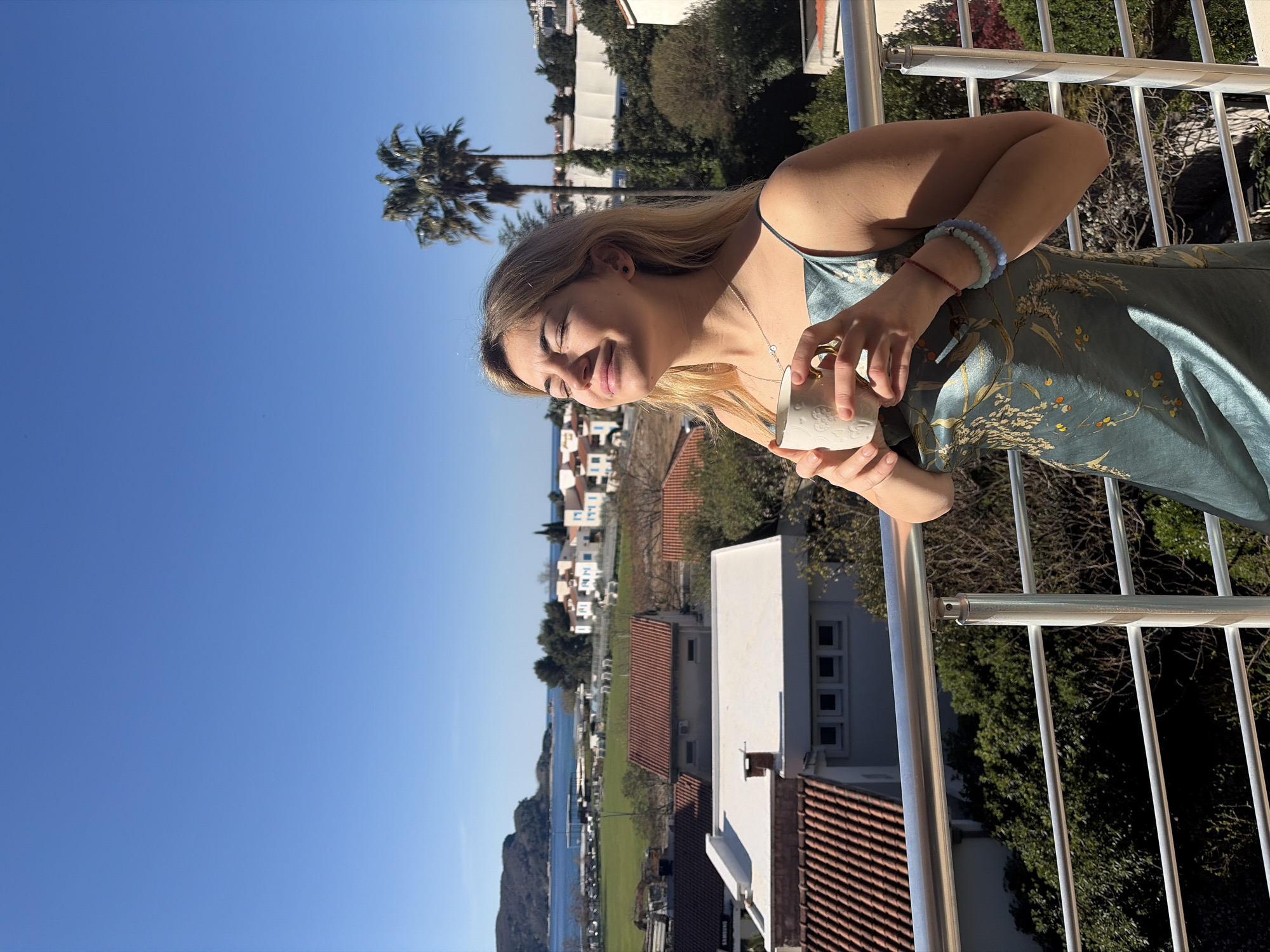 A woman with blonde hair and a blue dress stands on a balcony, holding a white mug. She is smiling. Behind her, the city of Budva, Montenegro, extends towards the sea. Palm trees are visible to the left. The day is sunny with a clear blue sky. Residential buildings with red-tiled roofs and green spaces are interspersed in the background.