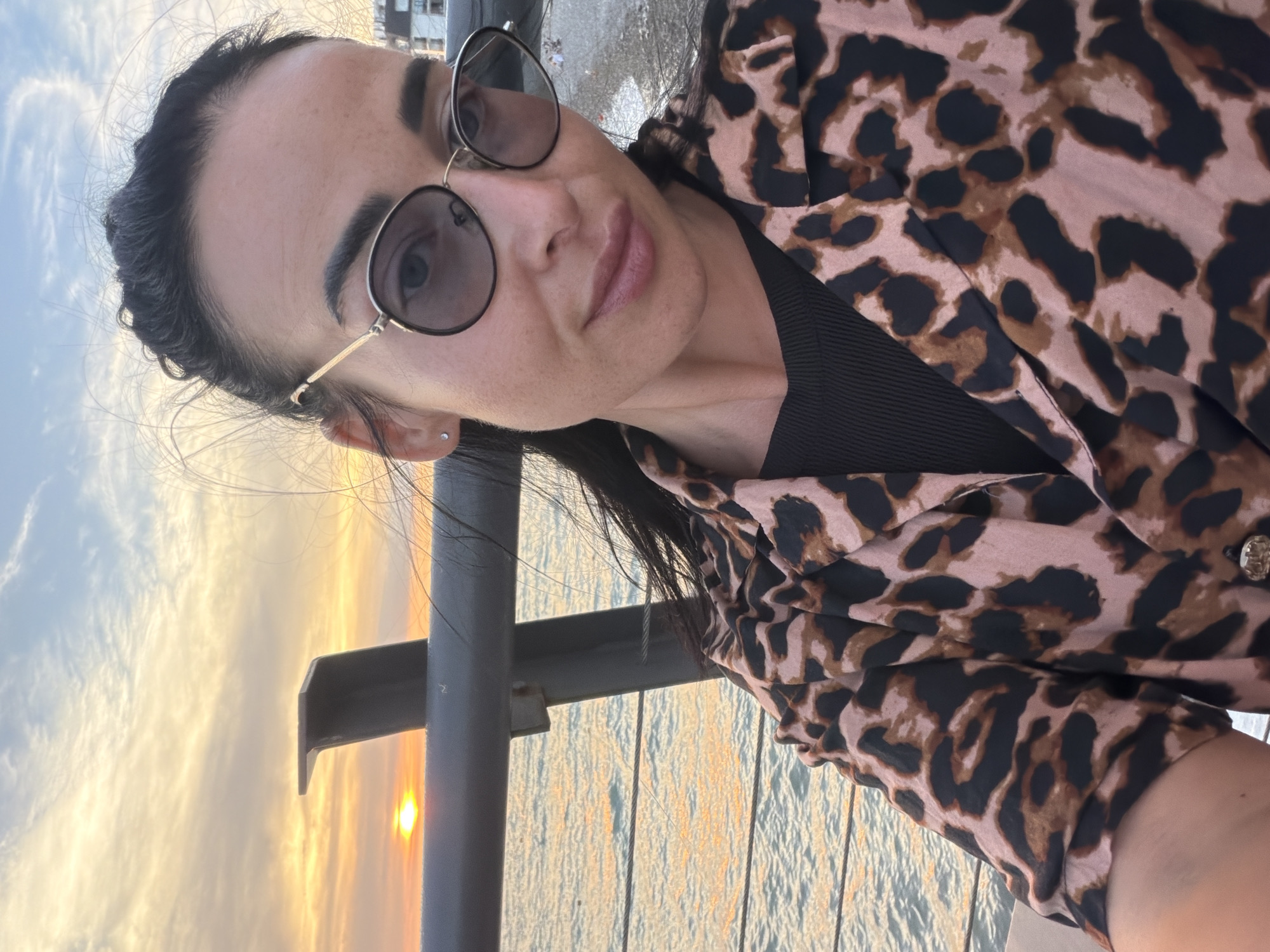 A woman with dark hair, wearing sunglasses and a leopard-print shirt, poses for a selfie on a pier at sunset in Sochi, Russia. The sun is setting over the Black Sea, casting a warm glow on the water and sky. A metal railing runs along the pier, and in the background, buildings can be seen along the coastline. The woman is smiling and looking at the camera.