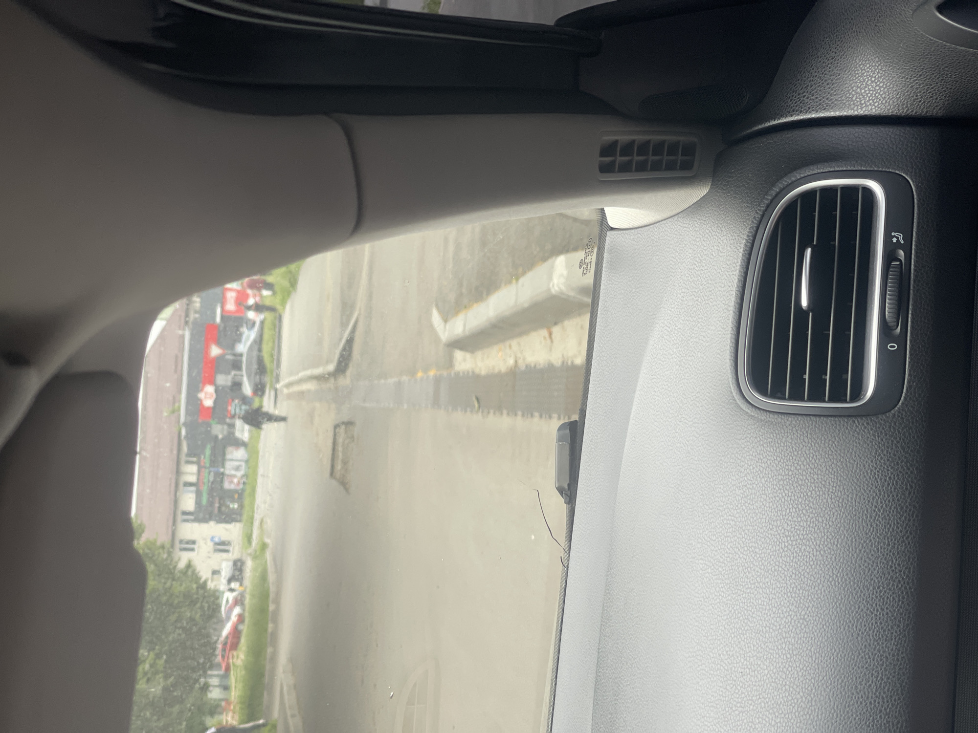 The image, taken from inside a vehicle, shows a street scene in what is identified as Гожули, Ukraine. The perspective is from the driver's or passenger's seat, looking forward through the windshield and partially obscured by the car's interior, including the dashboard and a sun visor.
The street appears to be a residential or commercial area with buildings visible in the background. There are several cars parked along the side of the road, and one red car is particularly noticeable. A white truck is also present. Two individuals are walking on the sidewalk or street. One person is wearing dark clothing and walking away from the camera. Another individual is partially visible in the distance. The road surface shows signs of wear, with visible potholes. A concrete curb and a drainage grate are also present along the side of the road. The sky is overcast, suggesting a cloudy day. The interior of the car shows a ventilation grate on the dashboard and a small crack on the windshield.