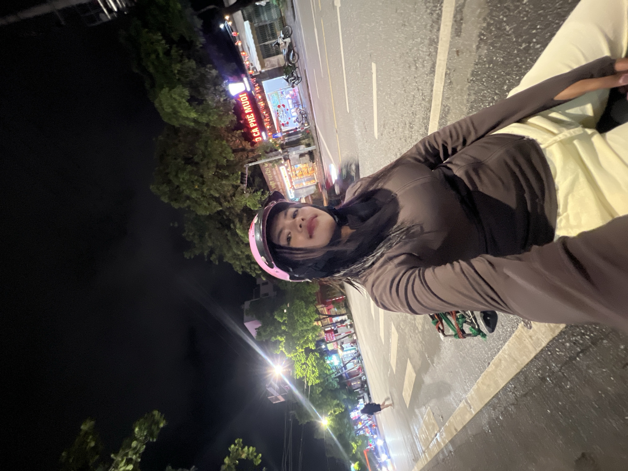 A young woman wearing a pink helmet and a brown jacket is taking a selfie on a motorcycle at night. The street is wet, suggesting it might have recently rained. In the background, there are shops and trees, with streetlights illuminating the scene. One shop sign in Vietnamese reads "BÒ CA PHE MUOI," indicating it is a coffee shop. Cars and motorcycles can be seen on the street. The woman is looking directly at the camera, with her hair wet.