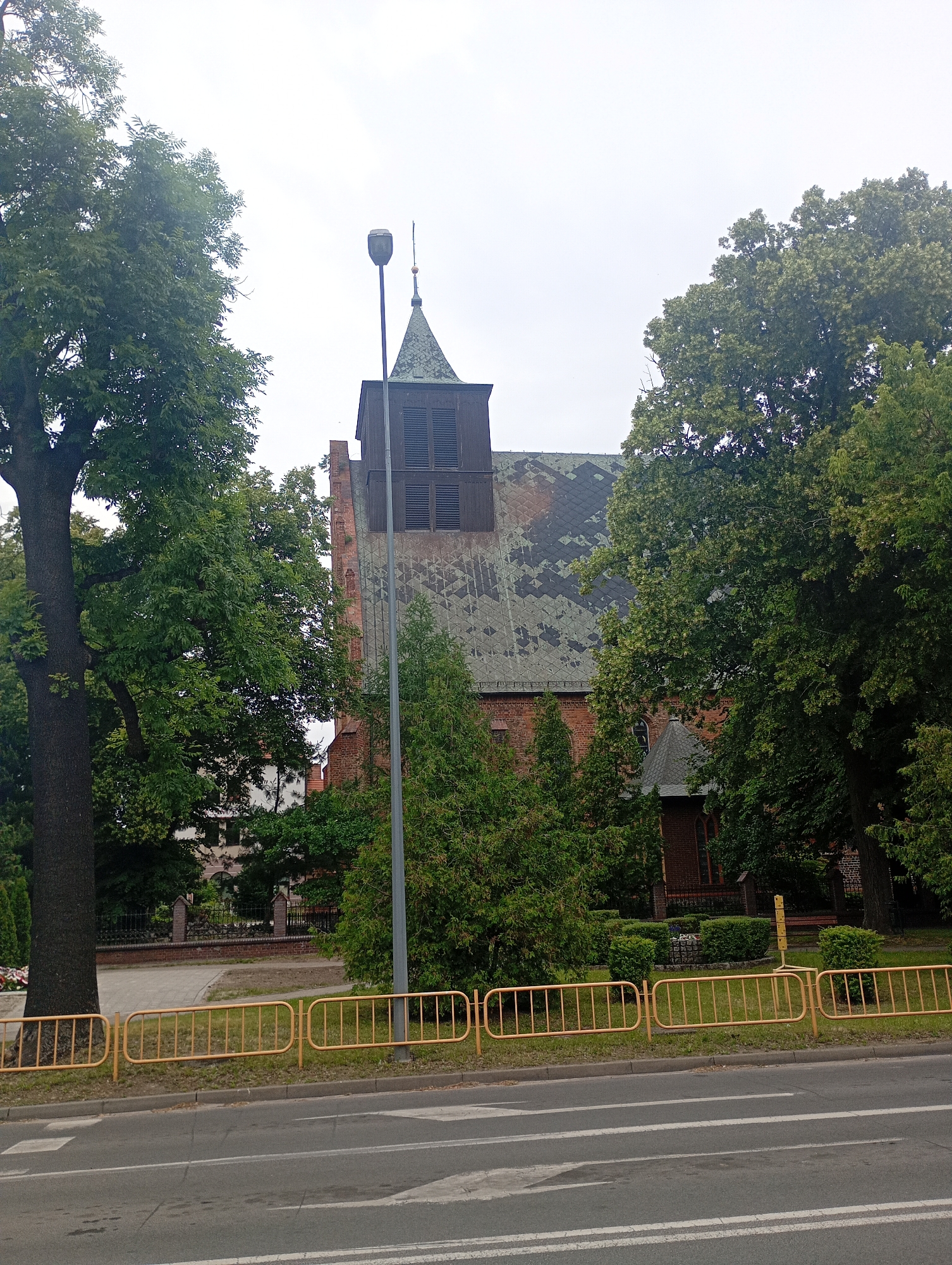 An outdoor, eye-level, medium shot captures a brick building with a dark, steepled roof adorned with a dark green, weathered material, likely shingles, showing some signs of discoloration and damage. The building features a tall, dark-wood tower with louvered openings, topped by a pointed spire. In front of the building, a light pole with a modern fixture stands tall. Lush green trees, some with thick trunks and expansive canopies, flank the building and the scene. A yellow metal barrier runs horizontally across the lower portion of the image, separating the building's grounds from a paved road with white lane markings. The sky above is overcast and a pale gray. The scene is set in the city of Międzyrzecz, Poland, specifically on ulica Wojska Polskiego, in front of the Church of St. John the Baptist.