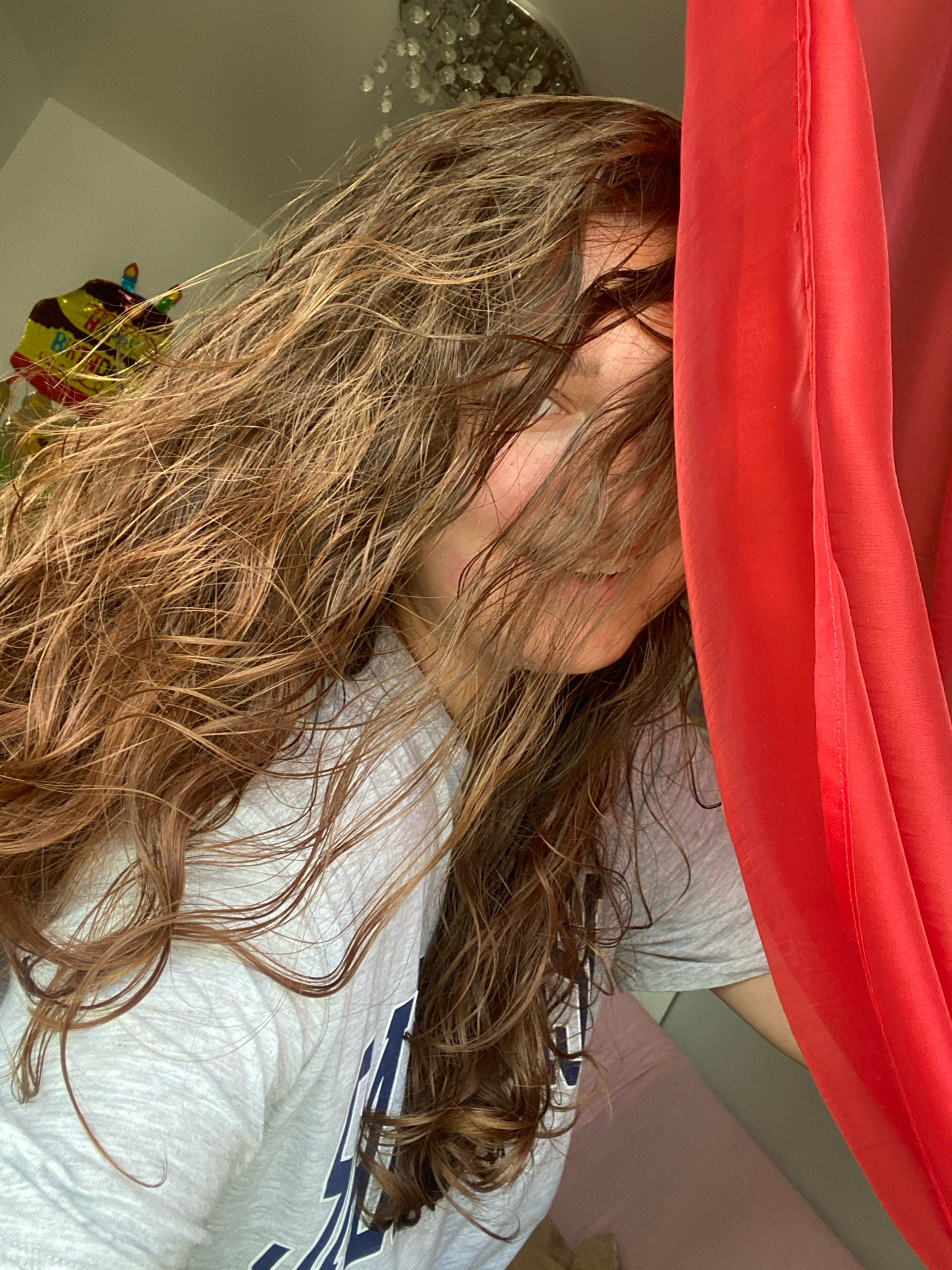 This is a medium shot, eye-level, indoor photograph of a young woman whose face is partially obscured by her long, wavy, brown hair and a red drape on the right side of the frame. She is looking towards the camera with a slight smile. Her hair is wet and appears to be styled in loose waves. She is wearing a light gray t-shirt with a navy blue design on the left side of the chest. In the background, on the left, a portion of a yellow and brown balloon shaped like a cake is visible, with a flickering light on top. Above the balloon and to the left, a portion of a ceiling fixture with dangling crystals is visible. The background is dimly lit. The location is an interior space, possibly a bedroom, in the city of Perm, Russia.