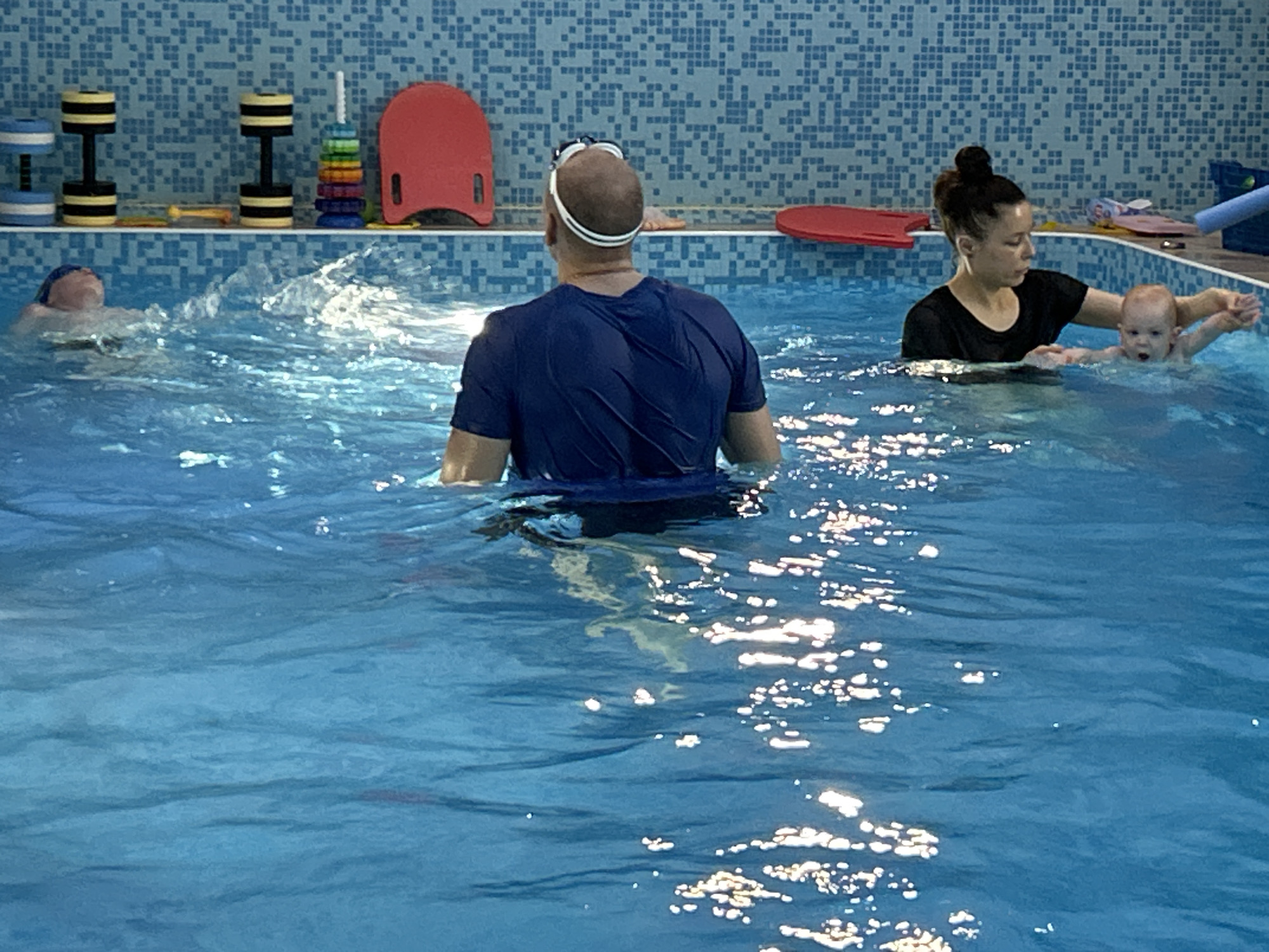 This image depicts an indoor swimming pool scene featuring four individuals. In the center foreground, a bald man, seen from behind, wears a blue short-sleeved shirt and has swimming goggles pushed onto his head. To his left, another person, wearing a blue swim cap, is in the water with their face upwards, creating a splash as they swim.
On the right side of the frame, a woman with her dark hair tied in a bun, dressed in a black short-sleeved top, is holding a baby in the pool. The baby, with light hair, has an open mouth, seemingly reacting to the water or the activity.
The pool water is a vibrant blue, showing ripples and bright reflections from overhead lighting. The background features a blue tiled wall, common in swimming facilities. Along this wall, a white shelf holds various swimming aids and toys. These include two pairs of water dumbbells (one blue and white, one yellow and black), a colorful stacking ring toy, and two red kickboards. A blue pool noodle fragment is also visible on the shelf. The scene suggests a swimming lesson or training session, possibly for young children or beginners, given the presence of the baby and the specific aquatic equipment. There is no visible text in the image.