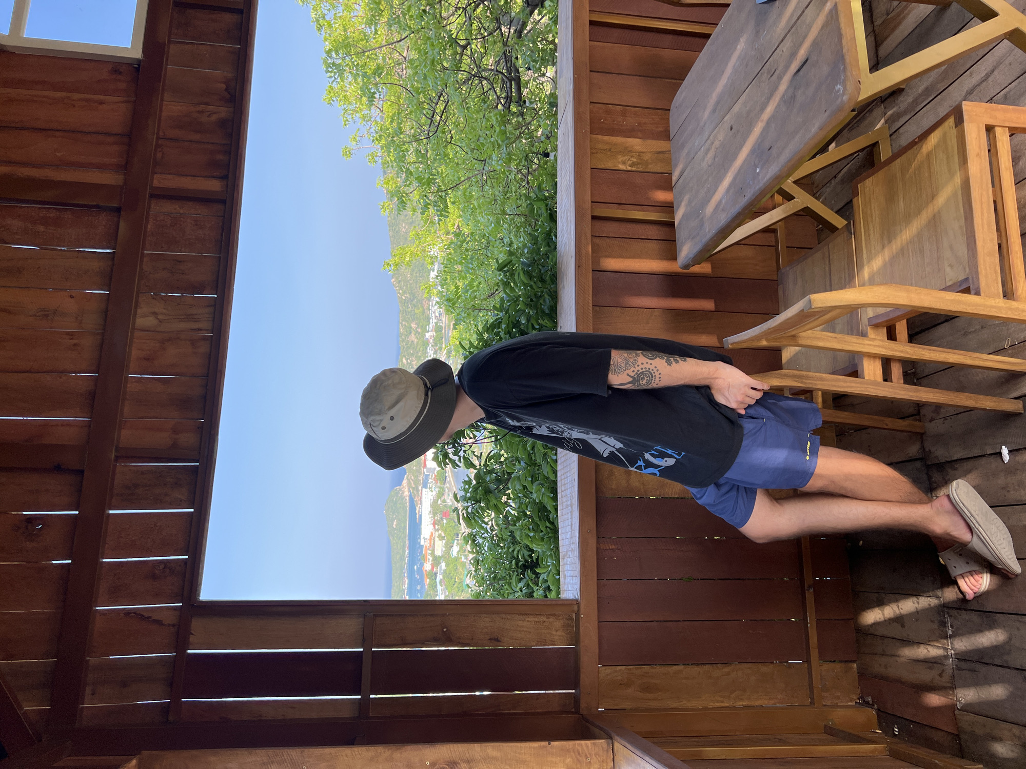 A male individual stands on a wooden observation deck or balcony, looking out at a scenic coastal view in Vĩnh Hải, Vietnam. The person is positioned in a three-quarter back profile, facing left, leaning against a sturdy wooden railing that forms the lower boundary of an open window-like aperture. He wears a dark, possibly black, graphic t-shirt with light-colored designs, navy blue athletic shorts, and light beige slip-on sandals. A beige and khaki bucket hat is on his head. A detailed, dark-inked tattoo featuring geometric and abstract patterns is visible on his left forearm.
The structure is constructed primarily from warm-toned wooden planks, forming the floor, walls, and ceiling. Gaps between the vertical planks on the walls allow slivers of light to penetrate. Sunlight creates distinct patterns on the wooden floor planks. To the right of the individual, a simple rectangular wooden table with a light-colored (possibly gold or light metal) X-frame base is accompanied by two matching wooden chairs, all appearing to be made of the same light-colored wood.
Beyond the wooden frame, a expansive, clear blue sky dominates the upper background. Below the sky, a panoramic view unfolds, featuring a vast body of blue water, likely a bay or ocean. An elongated island or peninsula, covered in dense green vegetation, is visible in the middle distance, extending into the water. Along the coastline, a compact town or city with numerous buildings, some with light-colored roofs, is nestled between the water and a verdant hillside. A road can be discerned winding through the town. In the immediate midground, lush green foliage and trees densely populate the terrain directly below the viewing platform. The scene is bathed in bright daylight, casting clear shadows and highlighting the vibrant colors of the landscape.