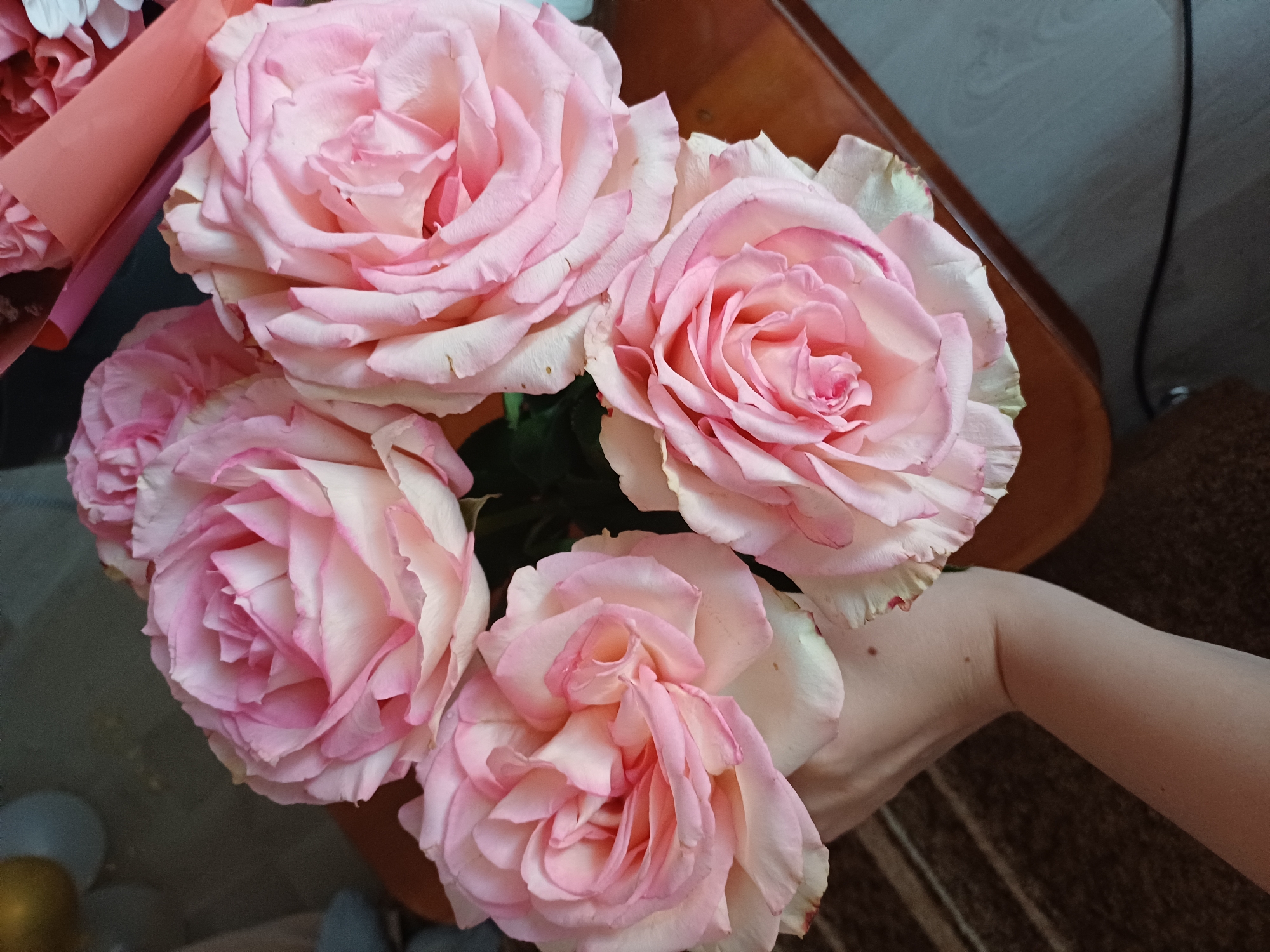The image shows a close-up of a bouquet of pink roses, held by a person's arm. The roses are the primary focus, with their delicate petals and soft pink hues filling most of the frame. The bouquet is partially wrapped in pink and orange paper. The background is blurred, but appears to be an indoor setting with a wooden surface and a dark, textured fabric. The lighting suggests it's daytime, possibly indoors with artificial lighting. There is no visible text in the image. The scene evokes a sense of softness and possibly a gift or decoration.