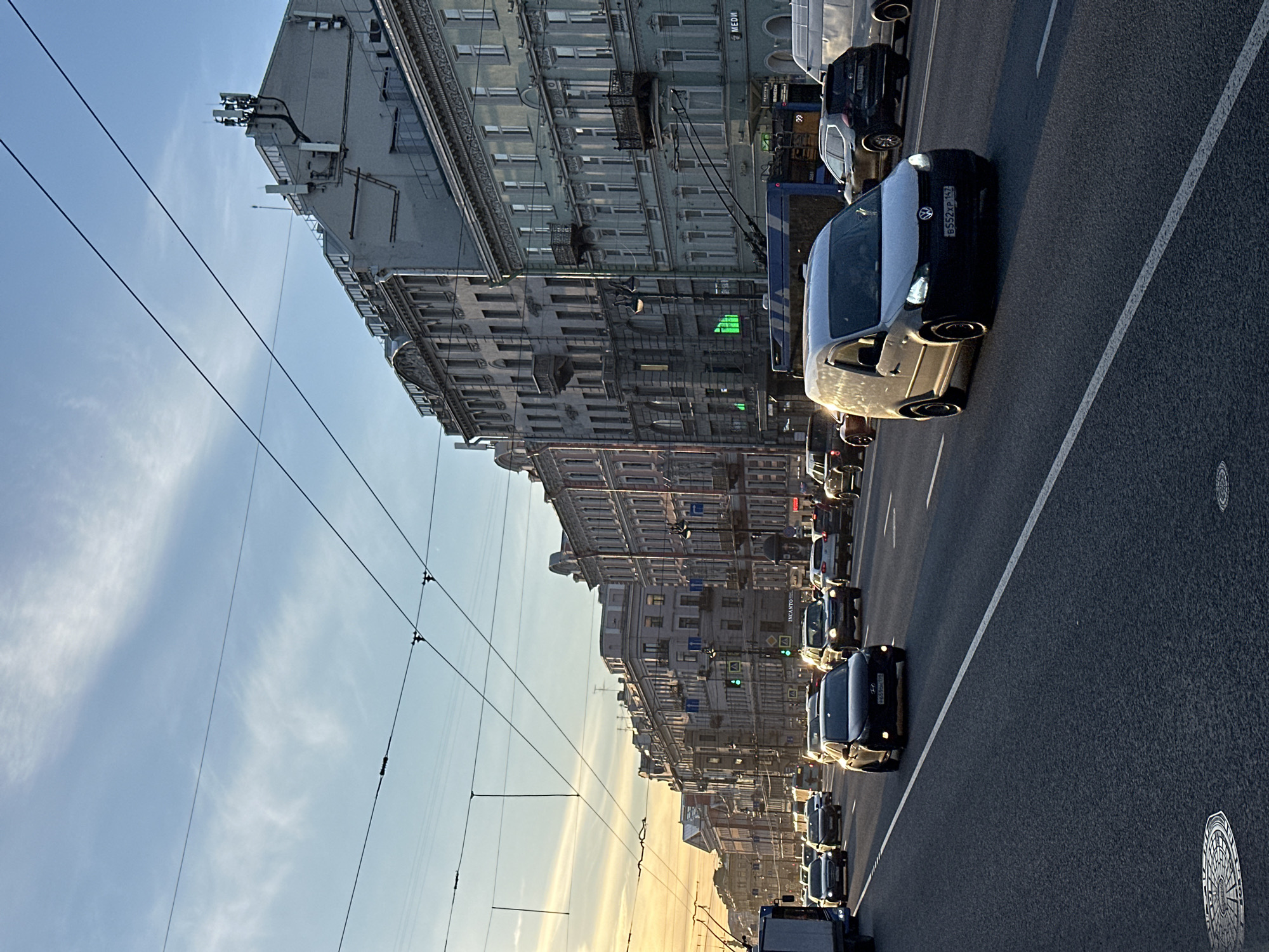 A street scene in Saint Petersburg, Russia, shows a line of vehicles on a multi-lane street bordered by tall buildings. The sky is partly cloudy with sunlight. Power lines are visible above the street. A white line marks the edge of the road, and a manhole cover is visible in the pavement. Street signs and advertisements are seen on the buildings, as well as traffic lights.