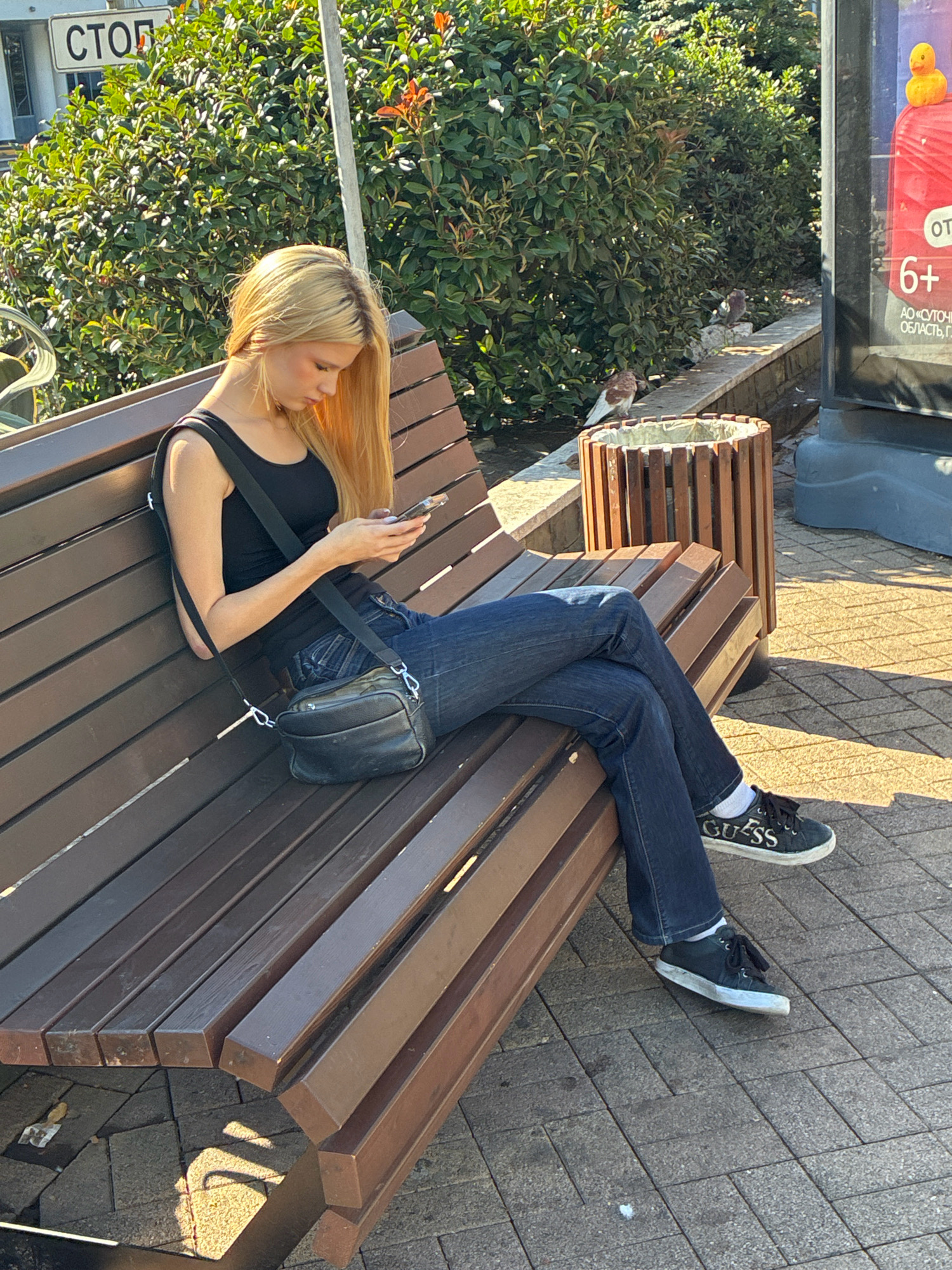 A young blonde woman with long hair, wearing a black tank top and blue jeans, is sitting on a wooden park bench in Sochi, Russia, and looking at her phone. She has a black crossbody bag slung over her shoulder. The bench is made of dark brown wood slats and is positioned on a paved walkway. Behind the bench is a dense green hedge, and a wooden trash receptacle. In the background, a billboard with a yellow rubber duck and the number "6+" is visible. The scene is illuminated by bright sunlight, casting shadows on the ground. Two pigeons are visible on a low stone wall near the trash receptacle. A white sign with the word "СТОП" (STOP in Russian) is attached to a pole behind the hedge.