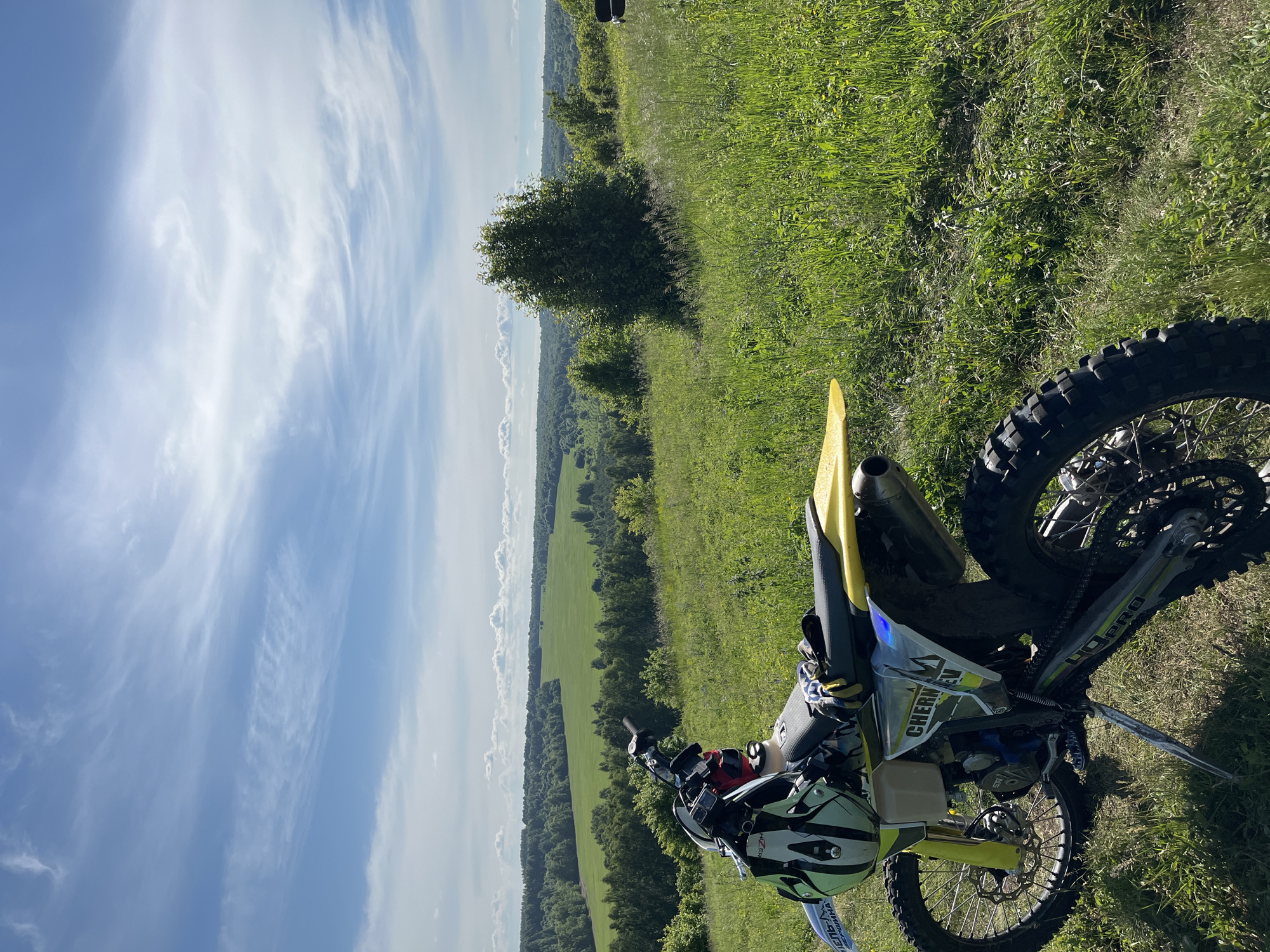 The image displays a rural outdoor scene in Vorob'evka, Russia, under clear visibility and natural illumination. In the foreground, a yellow, white, and lime green dirt bike is parked on its kickstand on a verdant, grassy hillside. An off-road helmet, predominantly light green and white with "HIZER" visible, rests on the handlebars/seat, alongside a pair of gloves. The bike features aggressive knobby tires, an exposed engine, and "CHERNIKOV" branding on its side panel. A faint "GoPro" logo is discernible on the swingarm. The mid-ground comprises the steep, grassy slope, characterized by tall weeds and a dense, dark green tree positioned to the right. Beyond this hillside, an expansive valley unfolds, showcasing large, cultivated green fields interspersed with extensive areas of dense forest. The horizon is marked by a distant tree line. The sky above is clear blue, dotted with scattered white cirrus and cumulus clouds. No human subjects are visible within the scene.
