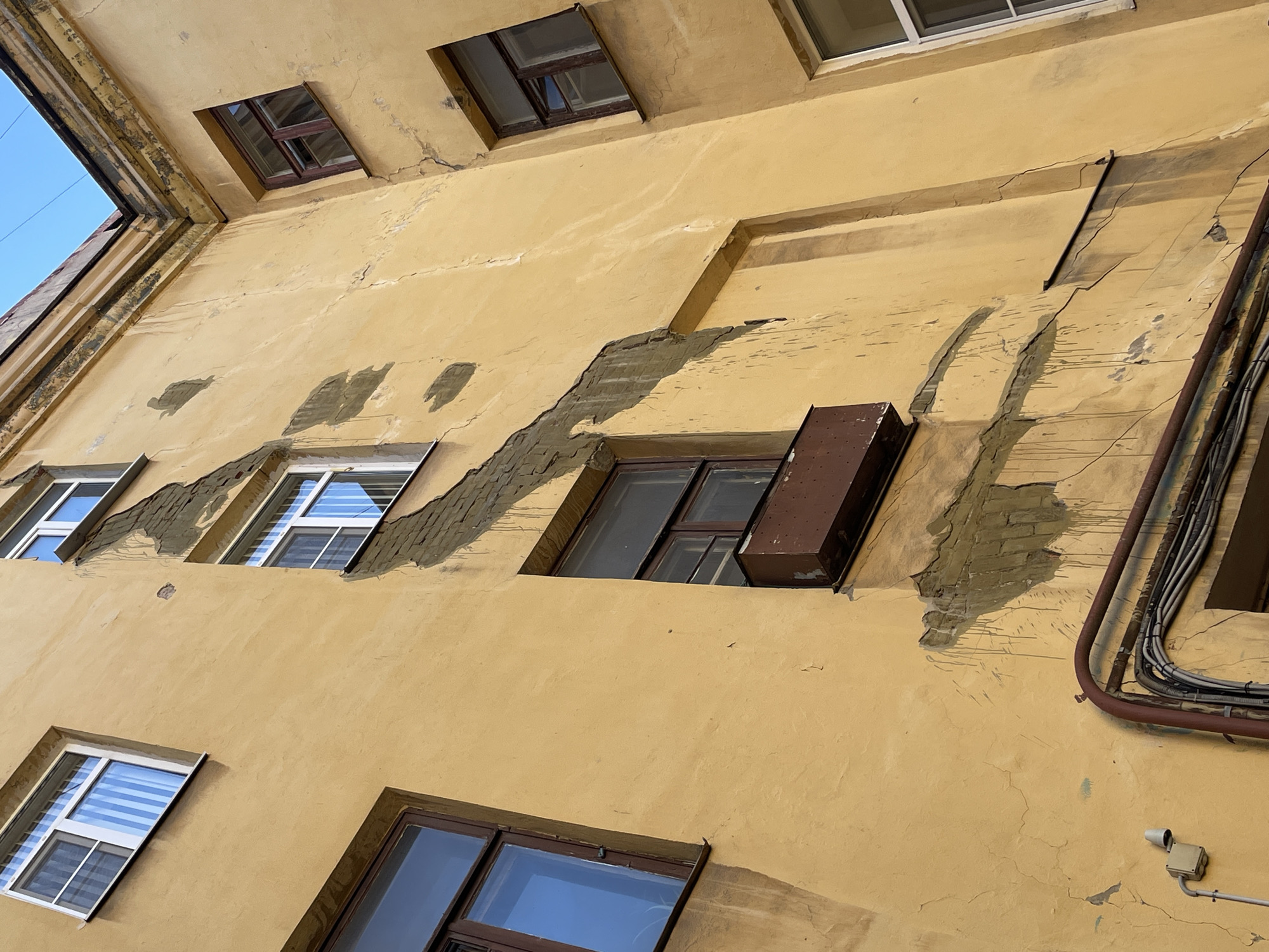 The image depicts the exterior wall of a multi-story building in Saint Petersburg, Russia. The wall is painted a faded yellow color and shows signs of significant wear and tear, including cracks, peeling plaster, and patches of exposed brick. Several windows are visible, some with wooden frames and others with white plastic frames. A metal planter box is attached to one window. Electrical conduits and cables run along the lower part of the wall, and a security camera is mounted on the left side. The architecture suggests an older building, possibly from the late 19th or early 20th century, common in the historic districts of Saint Petersburg. The sky is visible at the top of the image, indicating an outdoor scene.