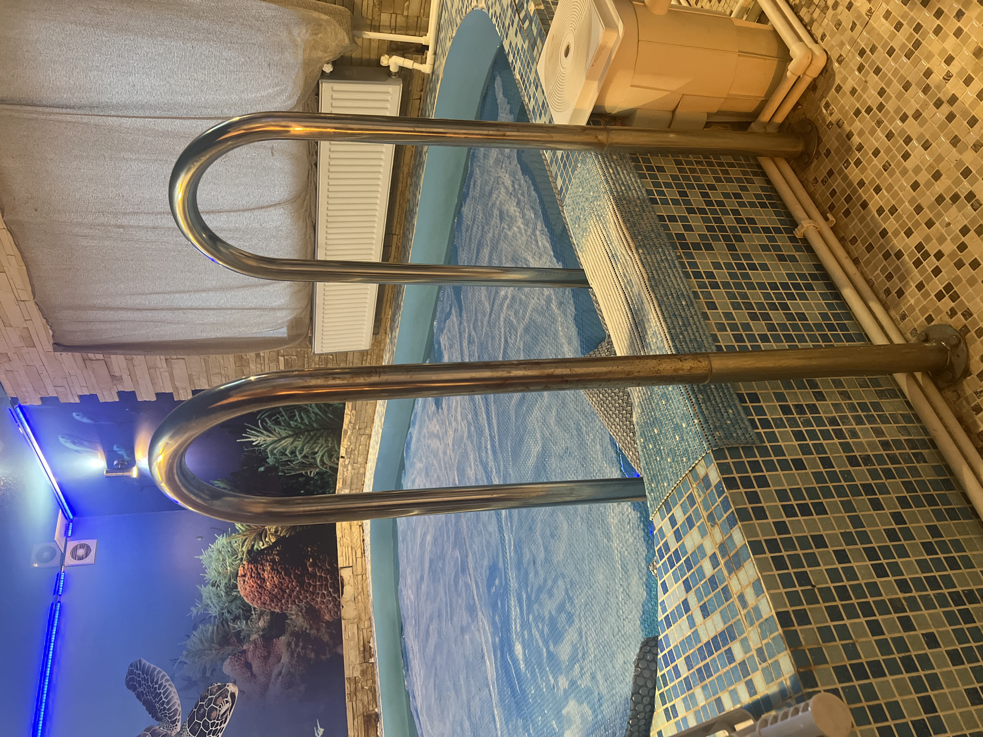 The image shows an indoor swimming pool in a building, possibly in Perm, Russia, but the exact location within Perm is unconfirmed. The pool is covered with a blue material, likely a pool cover. Two stainless steel grab rails are positioned at the edge of the pool for assistance entering and exiting the water. The pool's interior and surrounding area are tiled with blue and white mosaic tiles. One wall is decorated with a blue-toned mural depicting an underwater scene with marine life, including a sea turtle and coral. The opposite wall has beige and brown mosaic tiles with a heating radiator. A white filtration unit is positioned near the pool edge. The lighting conditions indicate artificial light is present, with blue LED strip lighting along the mural wall. No people are visible in the image.