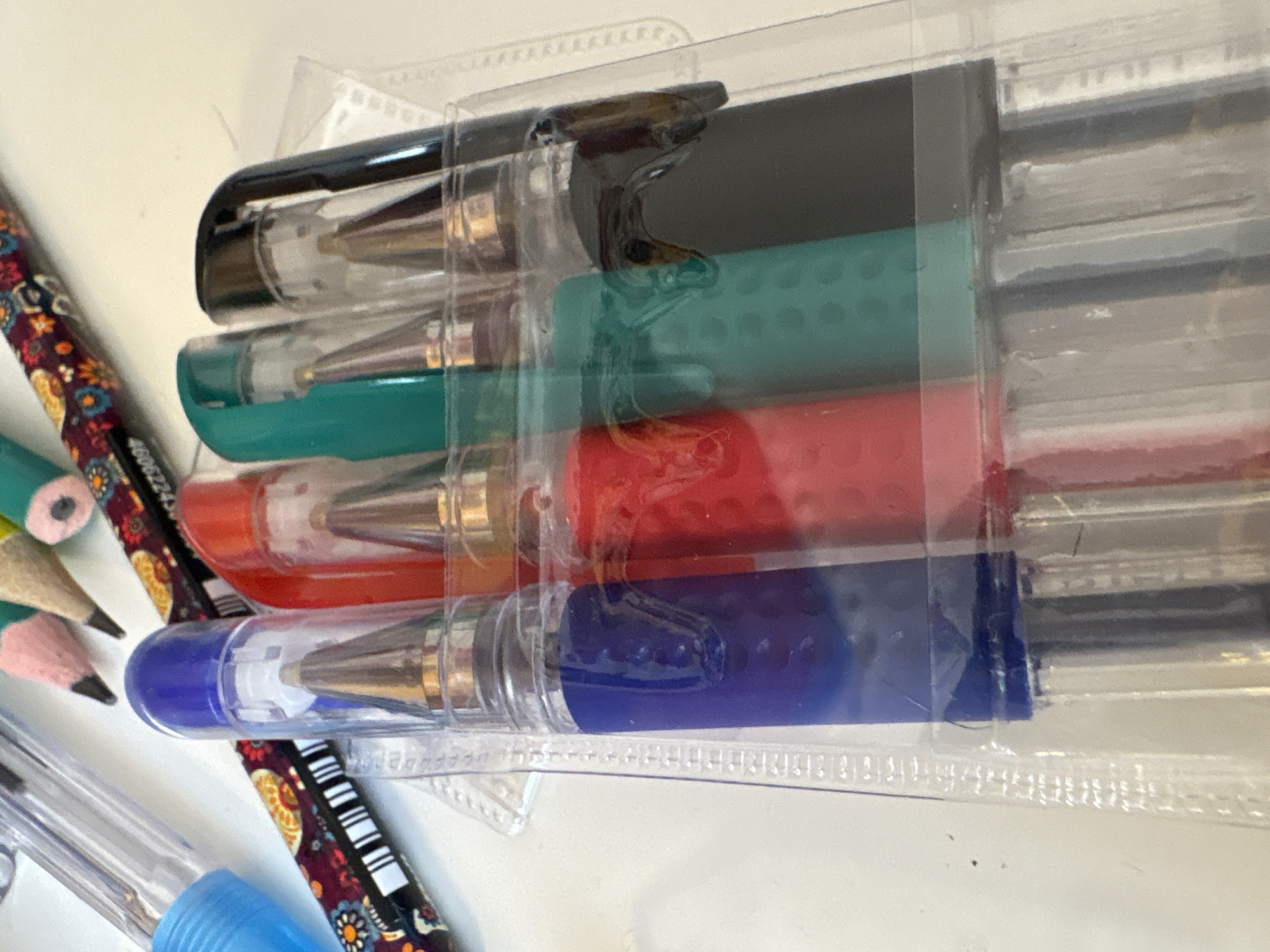 Four pens in blue, red, green, and black are arranged in a clear plastic case on a white surface. Other writing instruments, including pencils, are visible in the background. The image does not provide any information about the city of Georgiyevsk, Russia, or any other specific location.