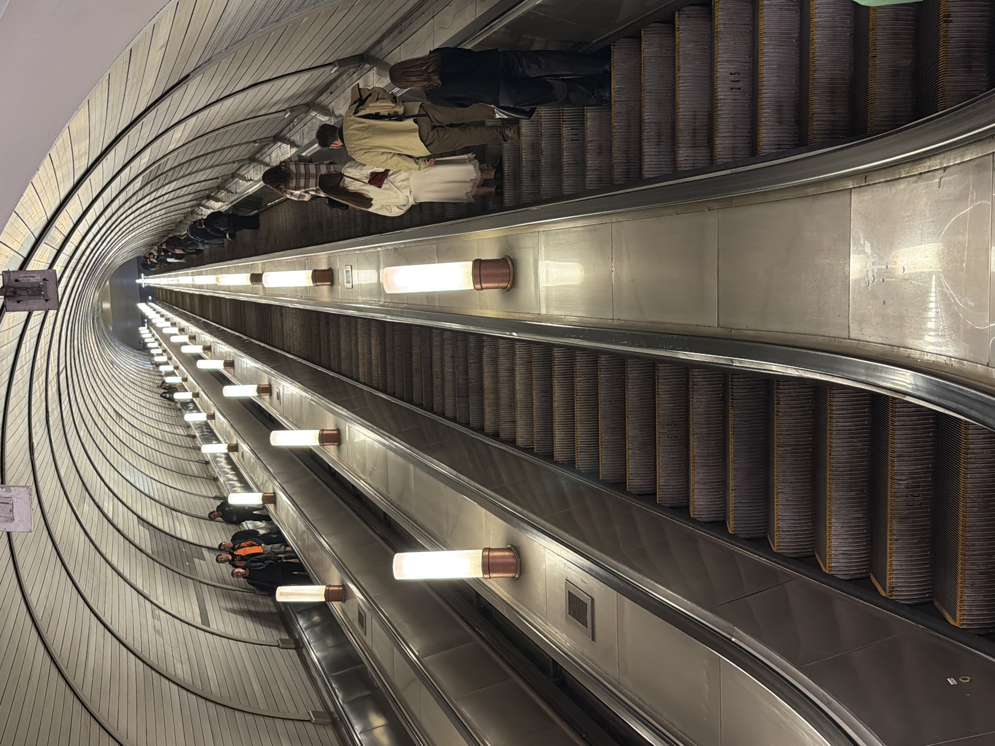 The image captures a long escalator descending into a subway station, possibly in an urban setting. People are riding the escalator, mostly facing downwards, suggesting they are traveling down into the station. The escalator has illuminated panels along the sides, and the station walls are curved and lined with metallic or light-colored panels, creating a tunnel-like effect. The overall scene gives a sense of depth and movement, as if the viewer is also descending into the subway system.