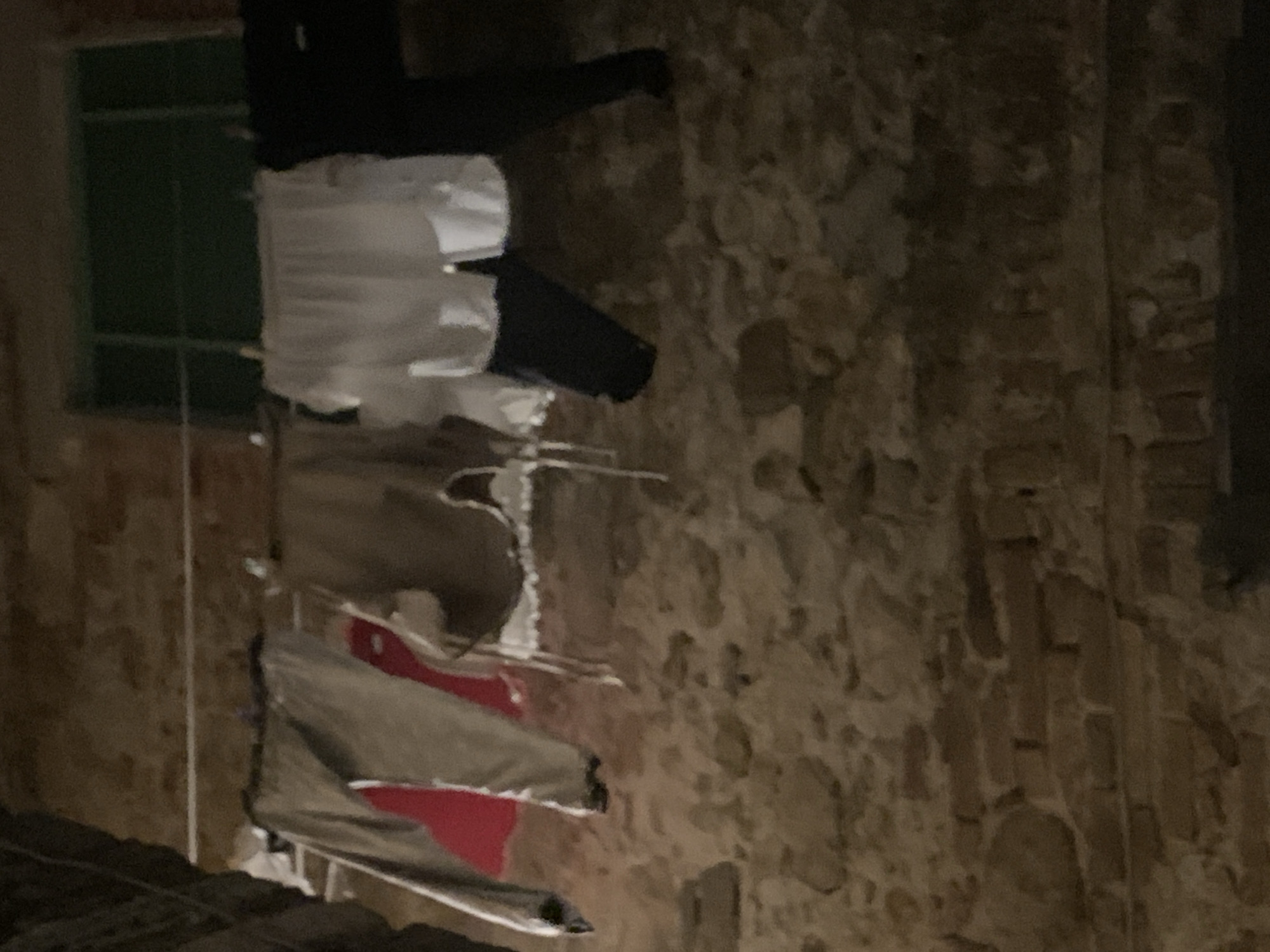 The image depicts laundry hanging on a clothesline in front of a stone wall, possibly in San Quirico d'Orcia, Italy. The laundry includes pants, a red garment, a brown garment, and white undergarments. A dark green window is visible in the background. The overall scene suggests a domestic setting, with clothes drying outdoors.