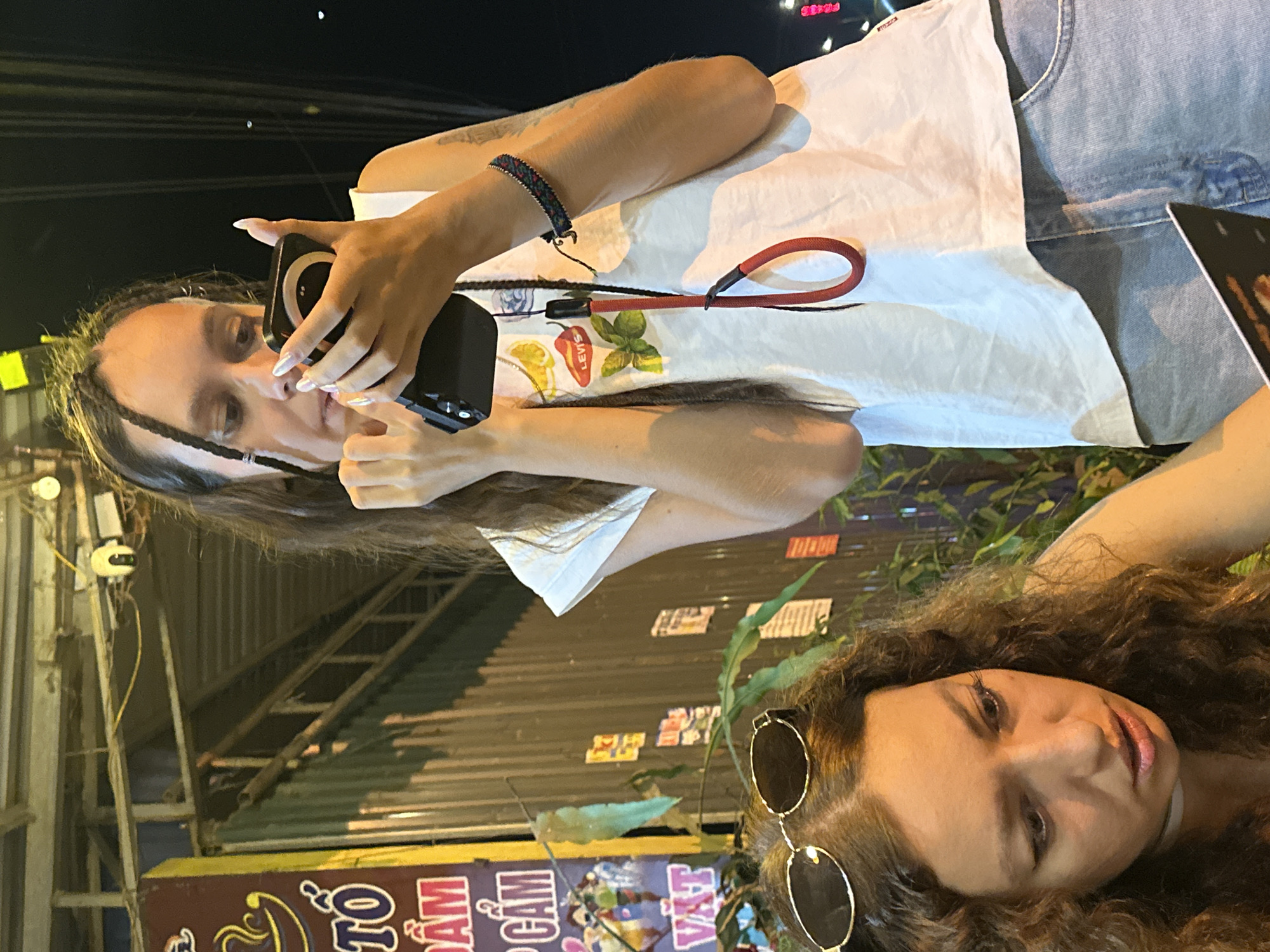 This image depicts two young women outdoors at night, likely in Mù Cang Chải, Vietnam. The scene is illuminated by artificial light, casting a warm glow on the subjects.
On the left, a woman with long, dark hair, featuring some braids, is holding a black smartphone equipped with a circular accessory on its back and a red lanyard. Her fingernails are light-colored and prominent. She wears a white t-shirt decorated with colorful patches of a lemon, a chili pepper, and a green leaf, with "LEVIS" text visible on the chili pepper patch. A multicolored woven bracelet adorns her right wrist. She appears focused on her phone, potentially taking a picture or recording.
On the right, partially visible, is another woman with voluminous, dark curly hair. She is looking slightly upwards and to the right with a soft, neutral expression. Round, gold-rimmed sunglasses are perched on top of her head.
The background consists of dark structures, appearing to be corrugated metal roofs or walls, typical of outdoor urban or semi-urban settings. Some green foliage and electrical wiring are also visible, along with a security camera mounted on a pole. At the bottom, a sign with Vietnamese characters, including "TÔ", "CẢM", and "VẬT", further establishes the location. The overall atmosphere is casual and relaxed.