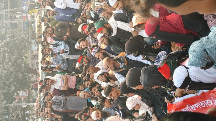 The image captures a large outdoor public gathering in the vicinity of the National Press Club, Dhaka, Bangladesh. The scene is densely populated with primarily young to middle-aged adults, interspersed with some children. Men stand in various parts of the crowd, many wearing traditional Islamic caps (taqiyah) and beards; some exhibit headbands featuring the green and red colors of the Bangladeshi flag. Women are predominantly seated in the immediate foreground, dressed in conservative Islamic attire, including hijabs and niqabs.
A red banner with white Bengali script, partially visible in the bottom-left, reads "...অধিকার বাঁচাও" ("...Save Rights"), indicating the event is a protest or rally. Several individuals, both standing and seated, are actively using mobile phones, likely for recording or photographing the event. In the mid-background, at least two men equipped with professional video cameras or stabilizers are observing, suggesting media coverage or organized documentation. The environment is an urban setting with a paved ground, numerous deciduous trees with sparse foliage, and indistinct multi-story buildings forming the background under hazy daylight conditions. A yellow auto-rickshaw or similar small vehicle is faintly visible behind the standing crowd.