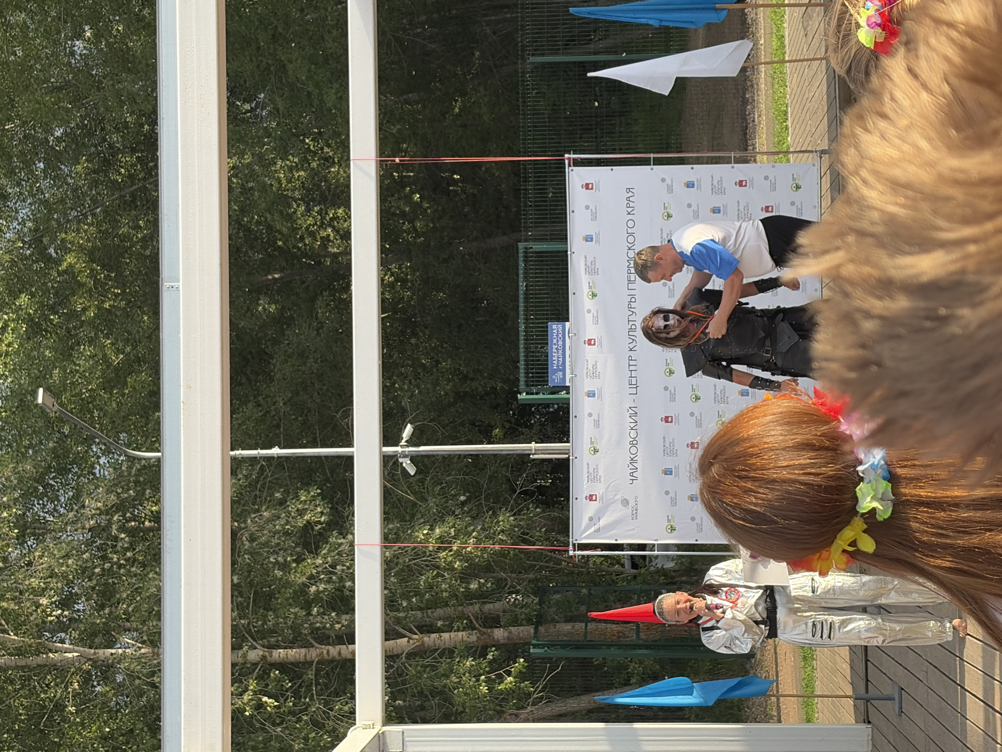 The image depicts an outdoor event on a wooden stage, likely in Chaikovsky, Russia, given the banner text. On the left, a woman in a silver astronaut-like costume with a red cone-shaped hat and a microphone is speaking. To her right, a person in a dark, medieval-style outfit with face paint resembling a zombie or a warrior stands. A man in a blue and white polo shirt is interacting with the costumed individual, appearing to adjust their attire. Behind them, a large white banner displays text in Russian, including "Чайковский - Центр Культуры Пермского Края" (Chaikovsky - Center of Culture of Perm Krai). Two flags, one blue and one red, are visible to the left of the stage. Another flag, white, and a blue umbrella or flag are visible to the right. In the foreground, the heads and hair of audience members are visible, one with a colorful lei around their neck. The background consists of trees and a fence. The lighting suggests it is daytime, with sunlight casting shadows.