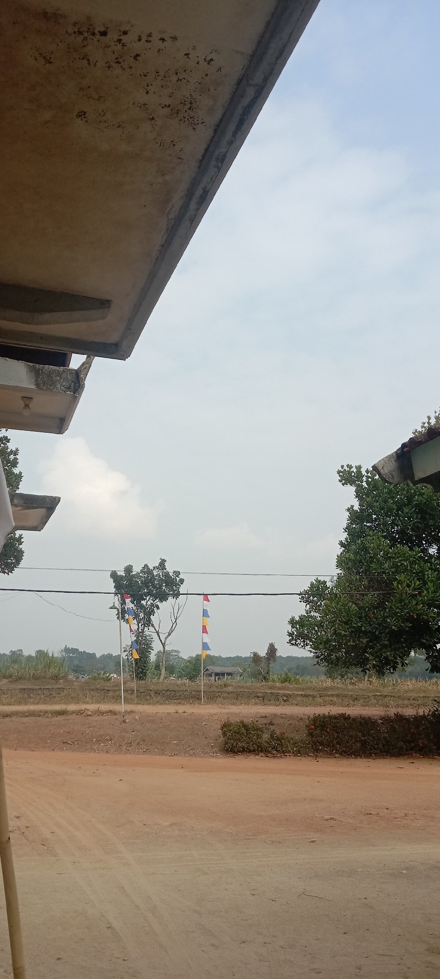 The image depicts an outdoor scene, likely in a rural or semi-rural area of Utama Jaya Mataram, Indonesia. In the foreground, a dirt path or road is visible with tire tracks. To the left, a portion of a concrete structure, possibly a building overhang or a roof, is present, showing signs of weathering and discoloration.
Further into the midground, a tree with bare branches stands to the left, near a pole with a string of colorful flags. These flags are rectangular and appear to be red, white, and blue. Another similar string of flags is visible to the right, attached to a pole near a large, leafy green tree. Between the trees, a simple building with a dark roof can be seen in the distance, surrounded by vegetation and what appears to be farmland or fields. Overhead, power lines stretch across the scene, and the sky is overcast with clouds. The overall environment suggests a quiet, open space.