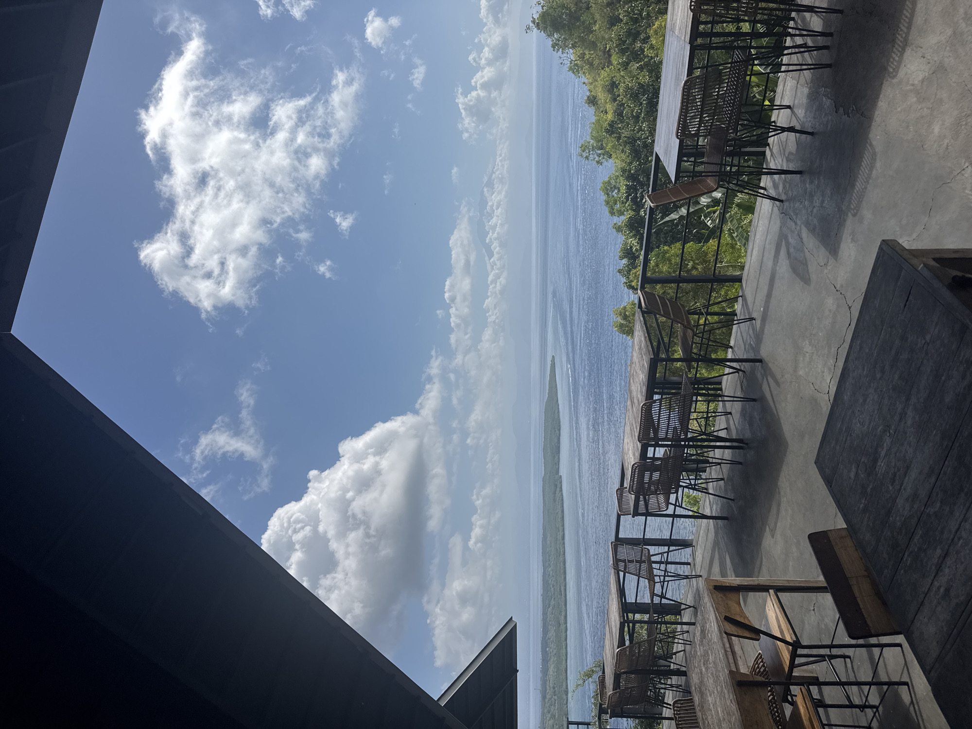 This image depicts an outdoor seating area with a scenic view of the ocean and islands. The seating area is located on a concrete patio with several wooden tables and wicker chairs. The patio is partially covered by a dark-colored roof structure. In the background, there is a body of water with islands and mountains visible in the distance. The sky is blue with scattered white clouds. The overall scene suggests a restaurant or cafe with an ocean view. This location is likely a popular spot for tourists and locals to enjoy the natural beauty of Nusa Penida, Indonesia.
