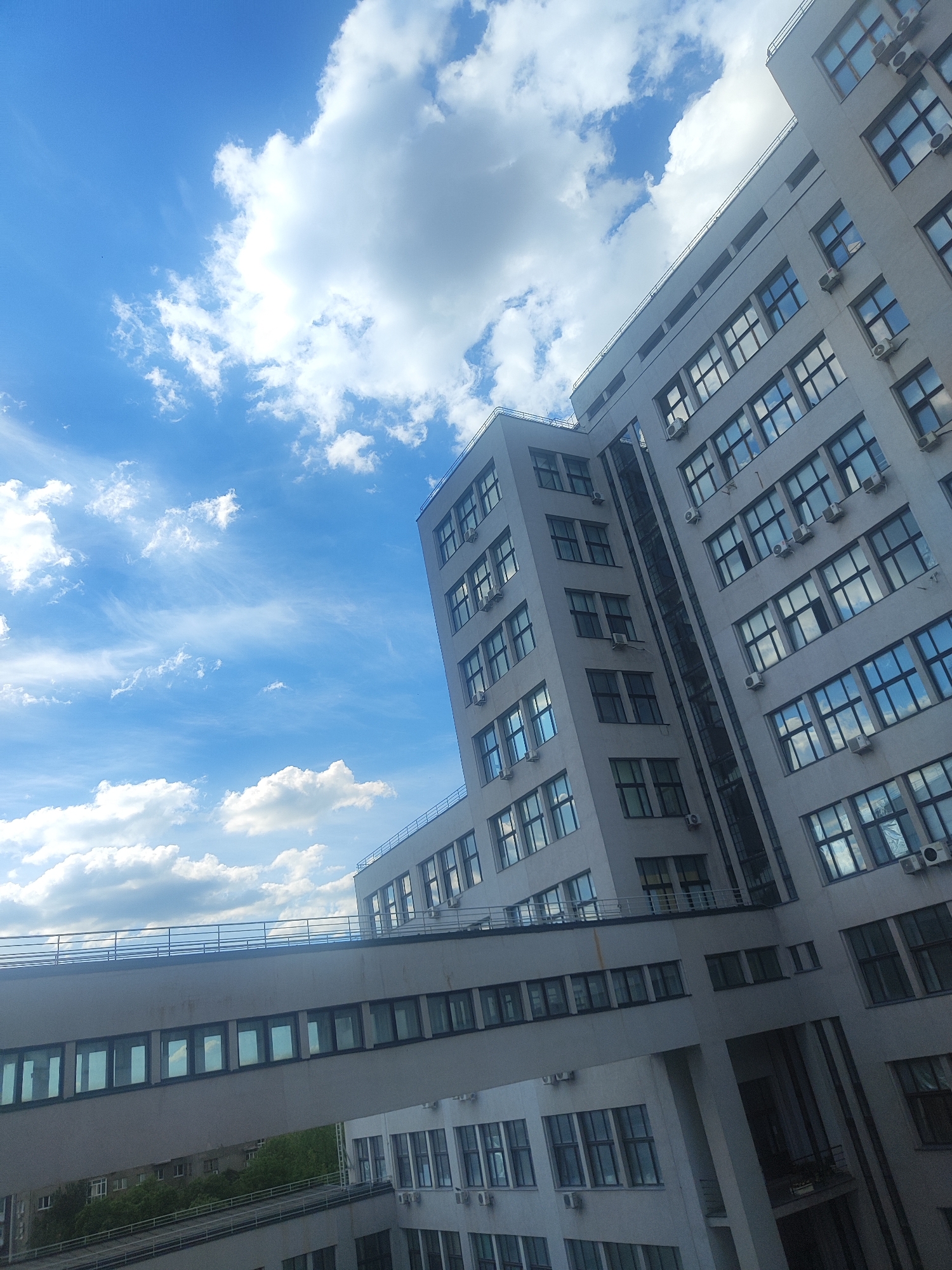 The image shows a modern, multi-story building with a skywalk connecting different sections. The building is made of light-colored concrete and features numerous windows, many of which reflect the blue sky and clouds. Air conditioning units are visible mounted on the exterior walls beneath some of the windows. A metal railing runs along the top of the building and along the skywalk. The sky is bright blue with scattered white clouds, suggesting it is daytime. In the background, glimpses of green trees and other buildings can be seen. The perspective of the photograph is angled upwards, emphasizing the height of the structure and the expanse of the sky.