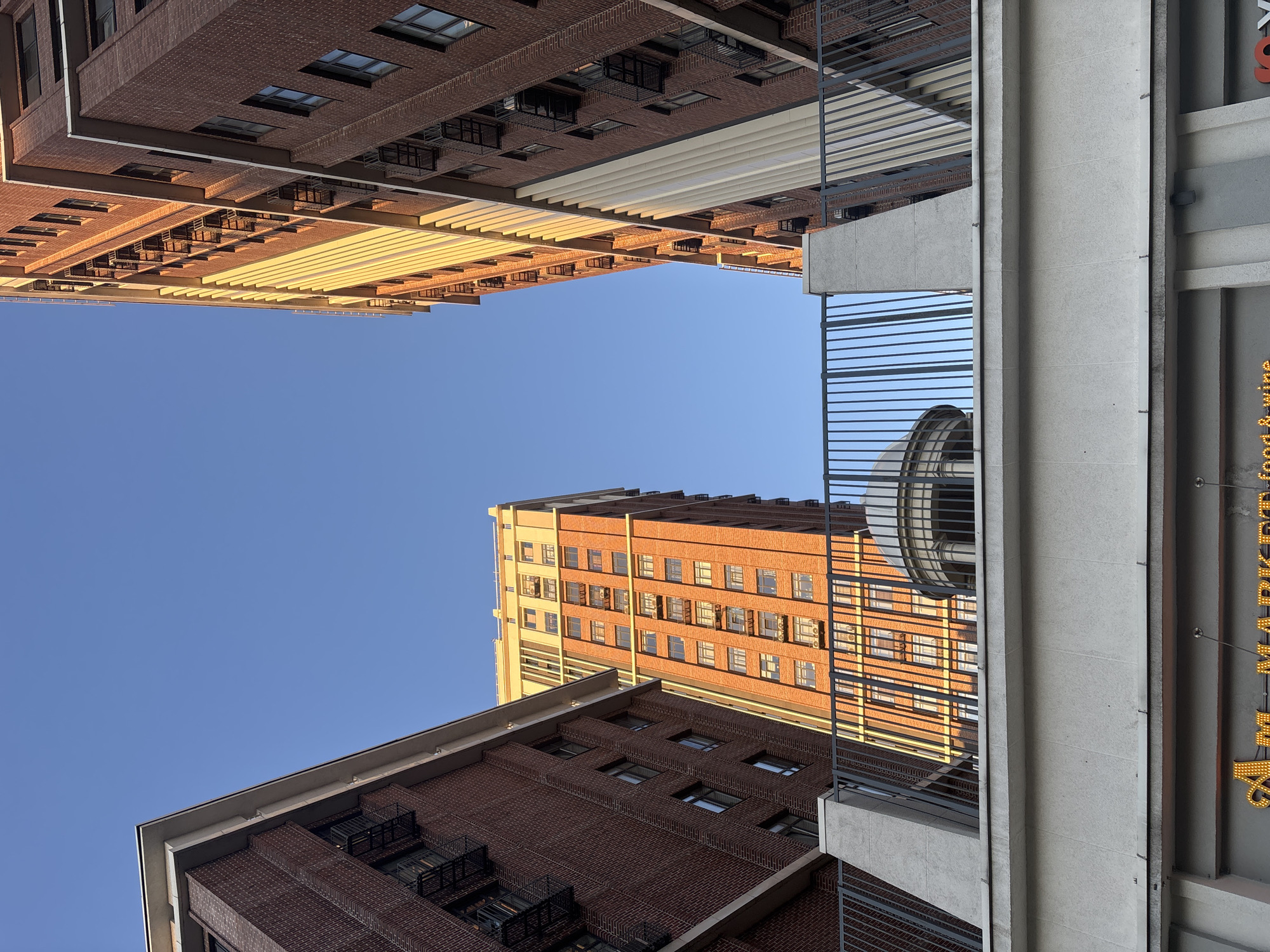 The image depicts an urban scene with tall brick buildings under a clear blue sky. Two prominent buildings, constructed with reddish-brown bricks, frame the view from the foreground. The building on the left has visible fire escapes and numerous windows. The building on the right, taller and illuminated by the warm glow of the sun, features a distinctive architectural element with vertical cream-colored fins. In the middle ground, a third, even taller building with a lighter facade and many windows is visible, also catching the sunlight. A grey metal fence with vertical bars runs along the top of a concrete structure in the foreground, partially obscuring a rounded domed structure behind it. Below the fence, a section of a building with the partial inscription "AM MARKET food & wine" is visible. The scene suggests a modern city environment, possibly a commercial or residential area, with a clear sky indicating fair weather. The perspective is from a lower vantage point, looking upwards at the architecture. This image does not show the city of Sinyavino, Russia.